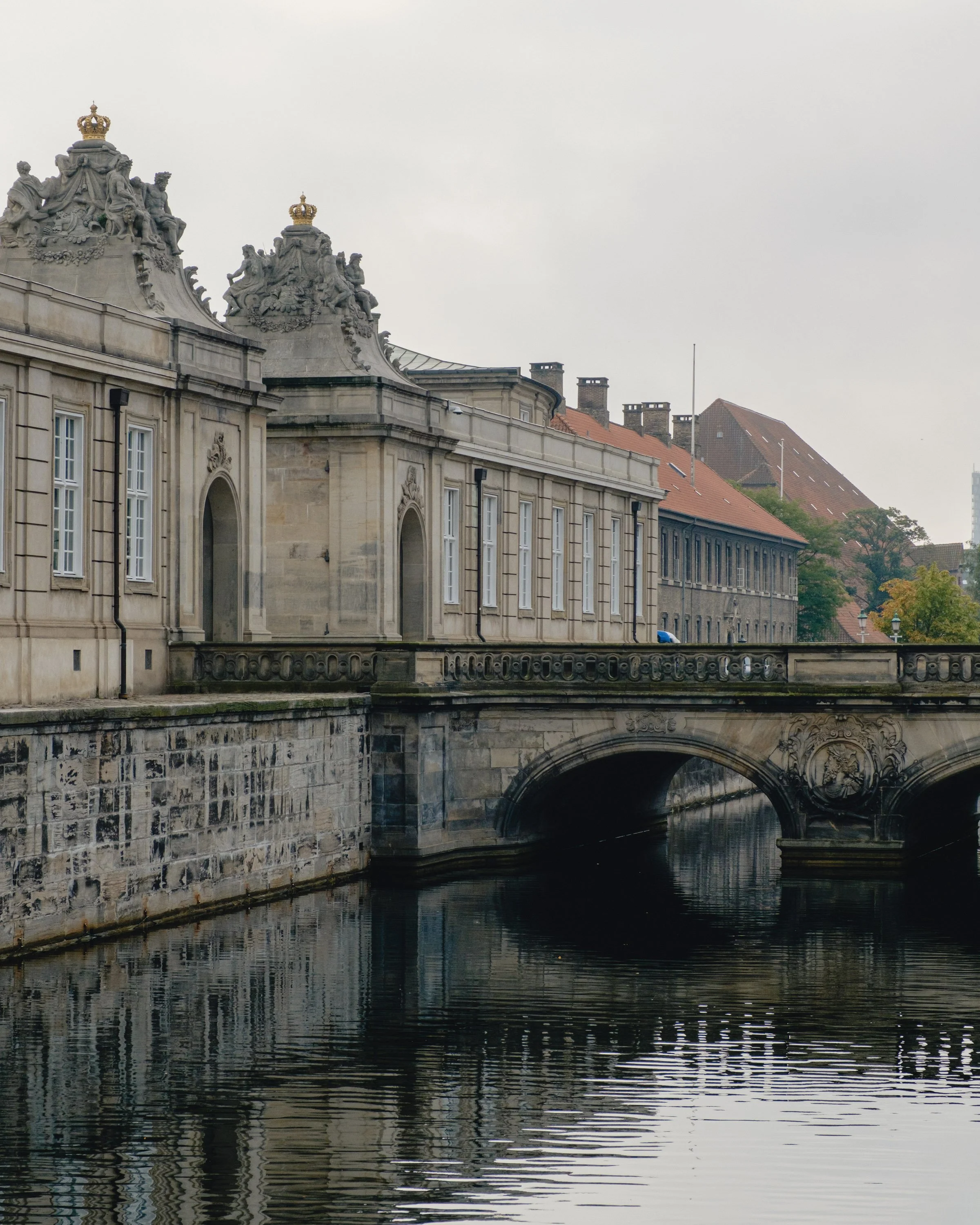 Historical building with ornate sculptures on its roof, a bridge over water, and old brick buildings with red roofs in the background.