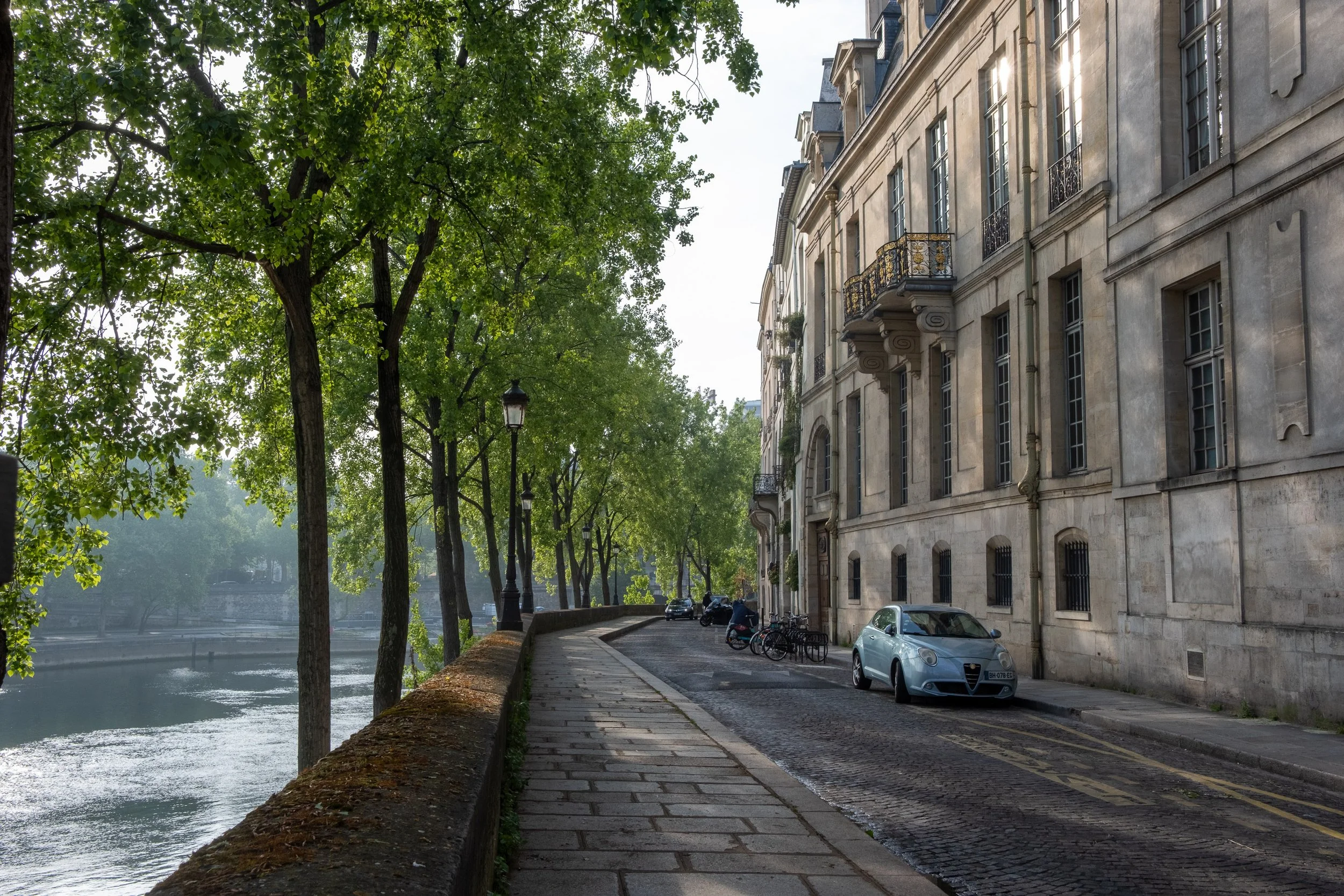 A quiet street beside a river with mature trees on the left and European-style buildings on the right, with parked cars and bicycles along the sidewalk.