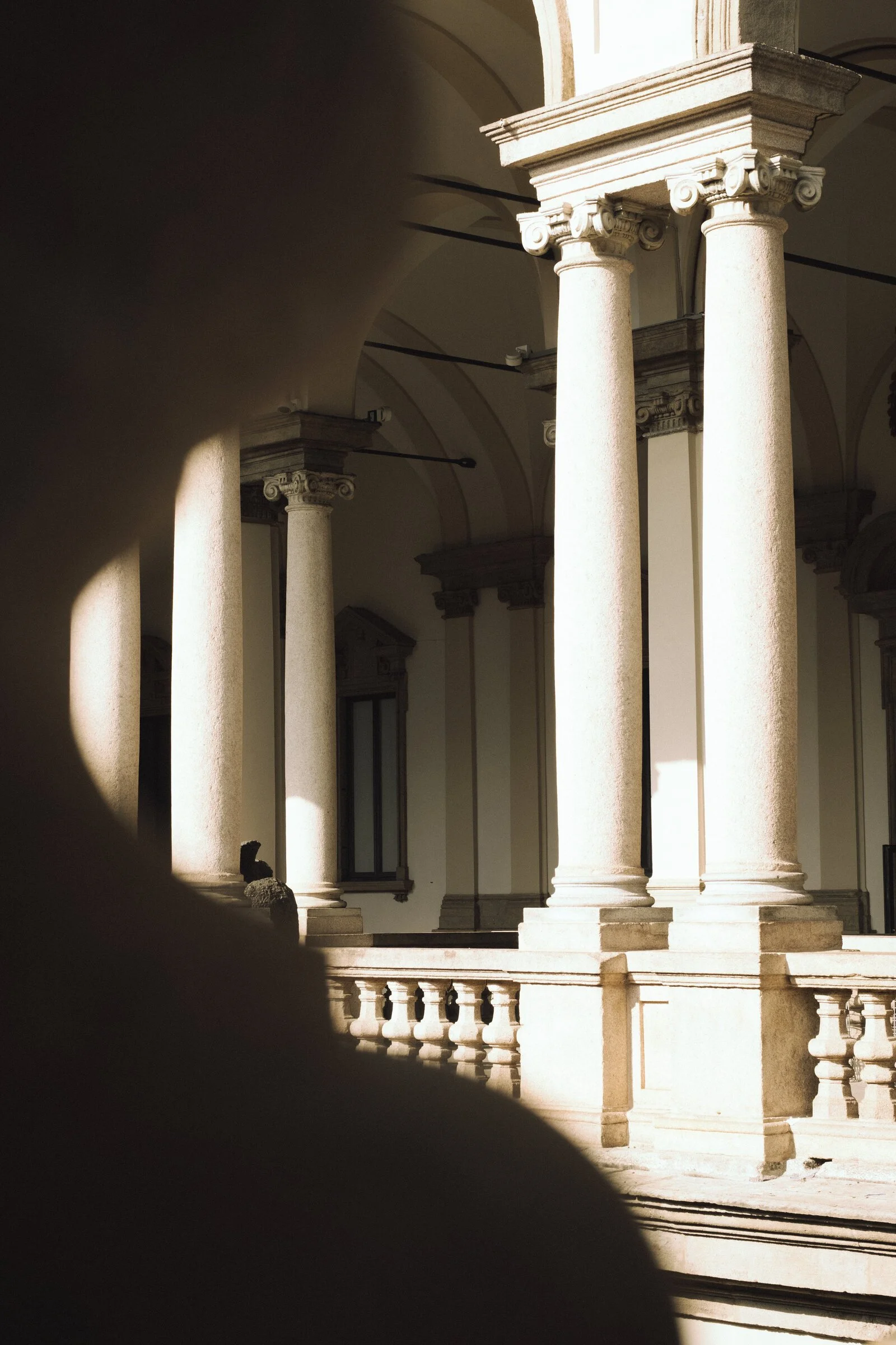 A sunlit stone colonnade in the Brera district, seen past the silhouette of a passerby's shoulder in the foreground