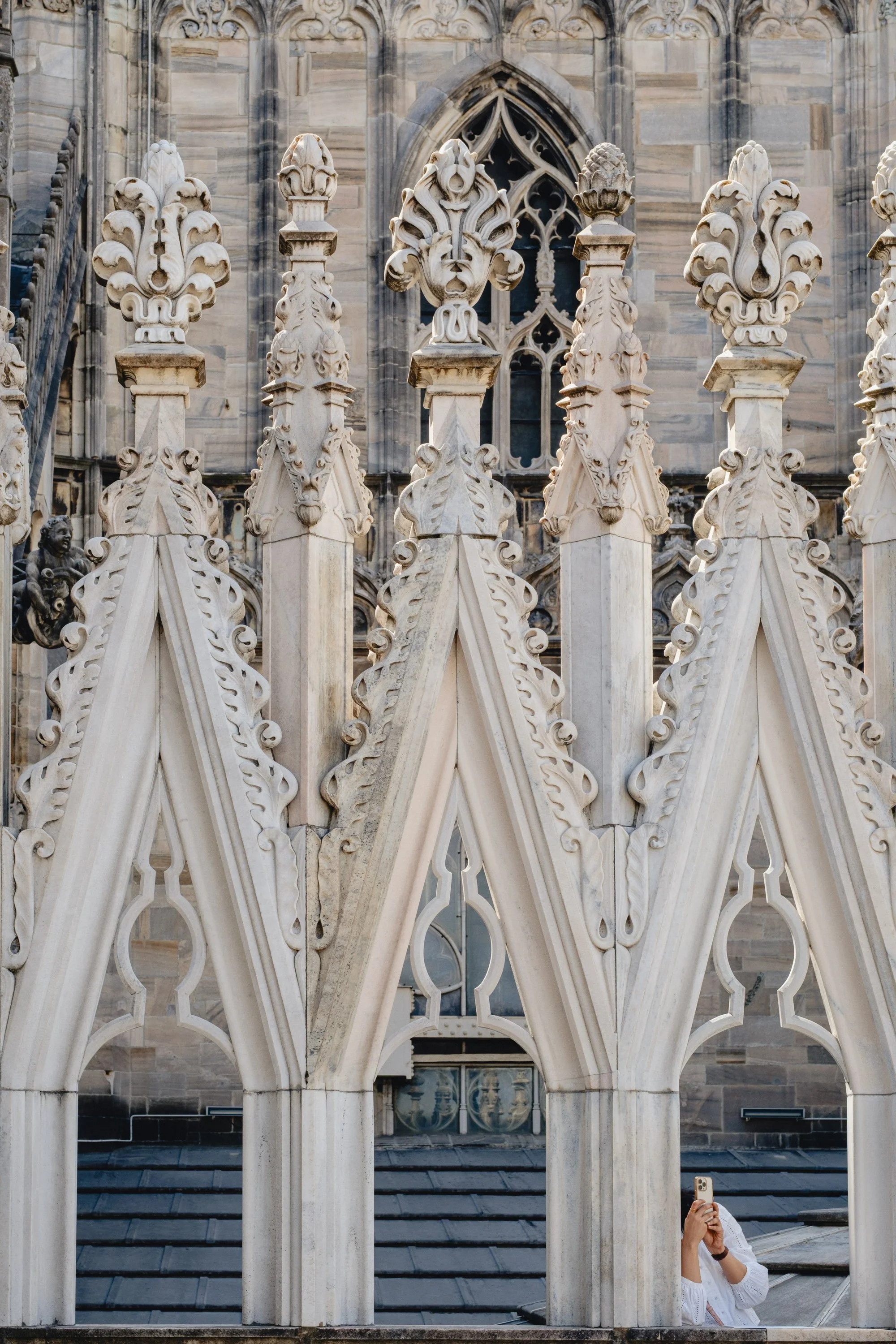 Close-up of ornate Gothic cathedral architectural details, including pointed arches and decorative stone carvings, with a person in a white top taking a photo with a smartphone in the lower right corner.