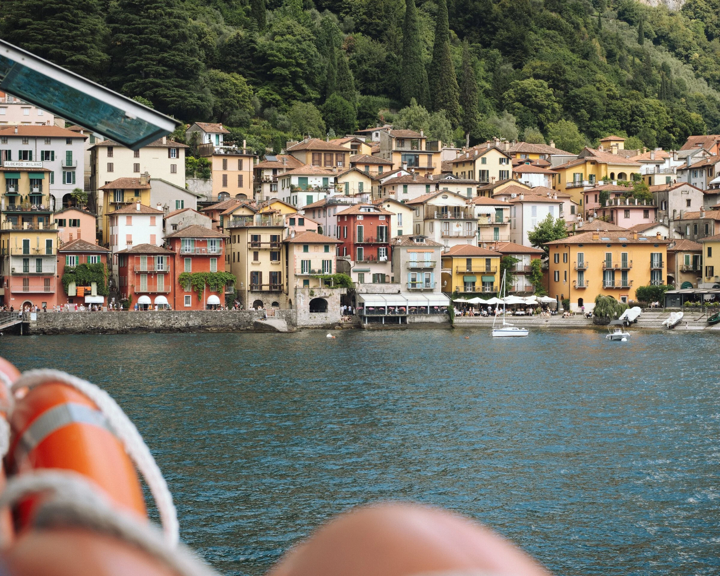 A colorful lakeside town's rooftops seen from a boat, framed by an orange life ring and a passenger's hand in the foreground