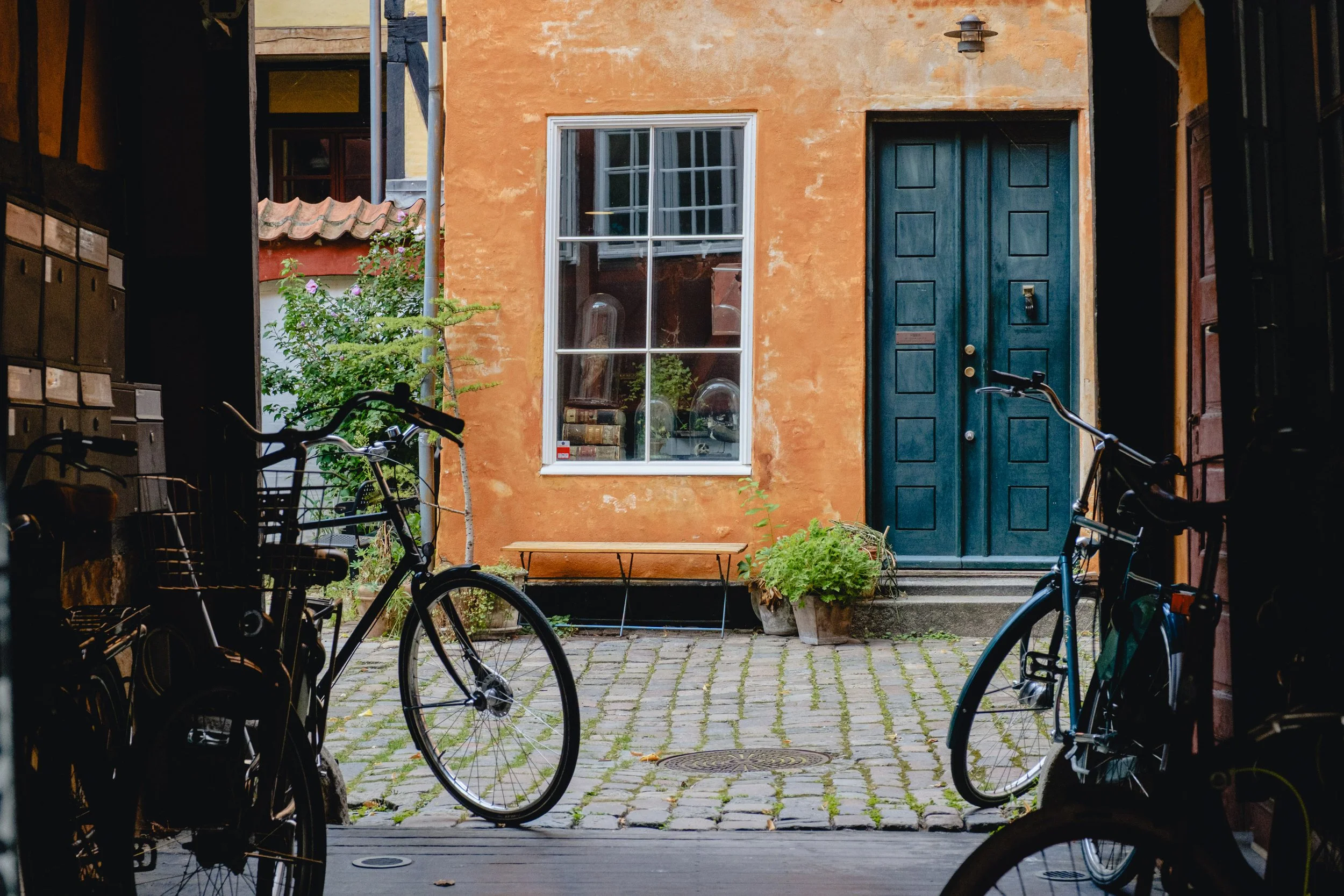 View of a cozy courtyard with a cobblestone floor, orange wall with a large window, green plants in pots, and a blue front door. Two bicycles are parked in the foreground.