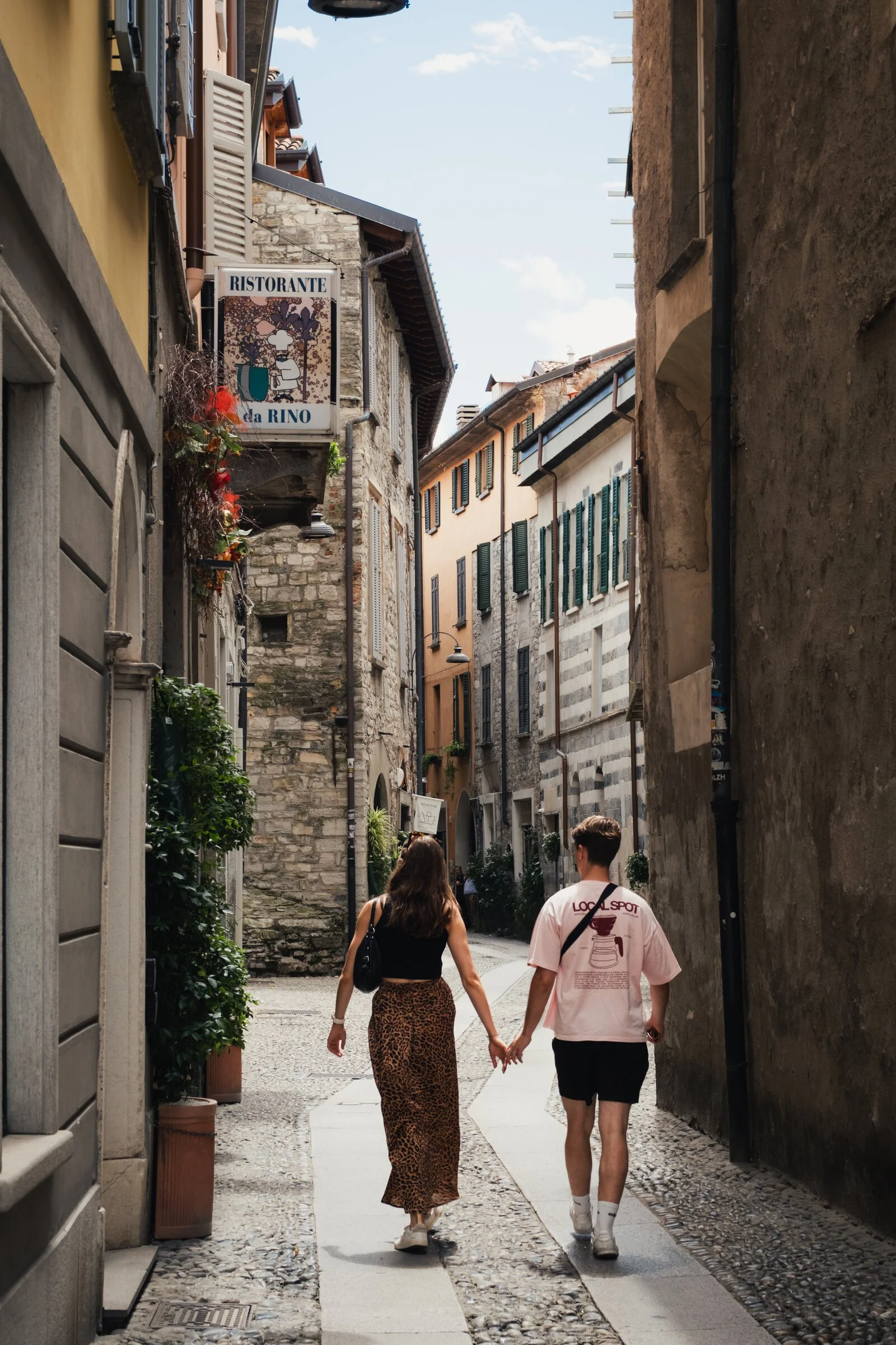 A couple walks hand in hand down a narrow cobbled alley past Ristorante da Rino in a Lake Como village