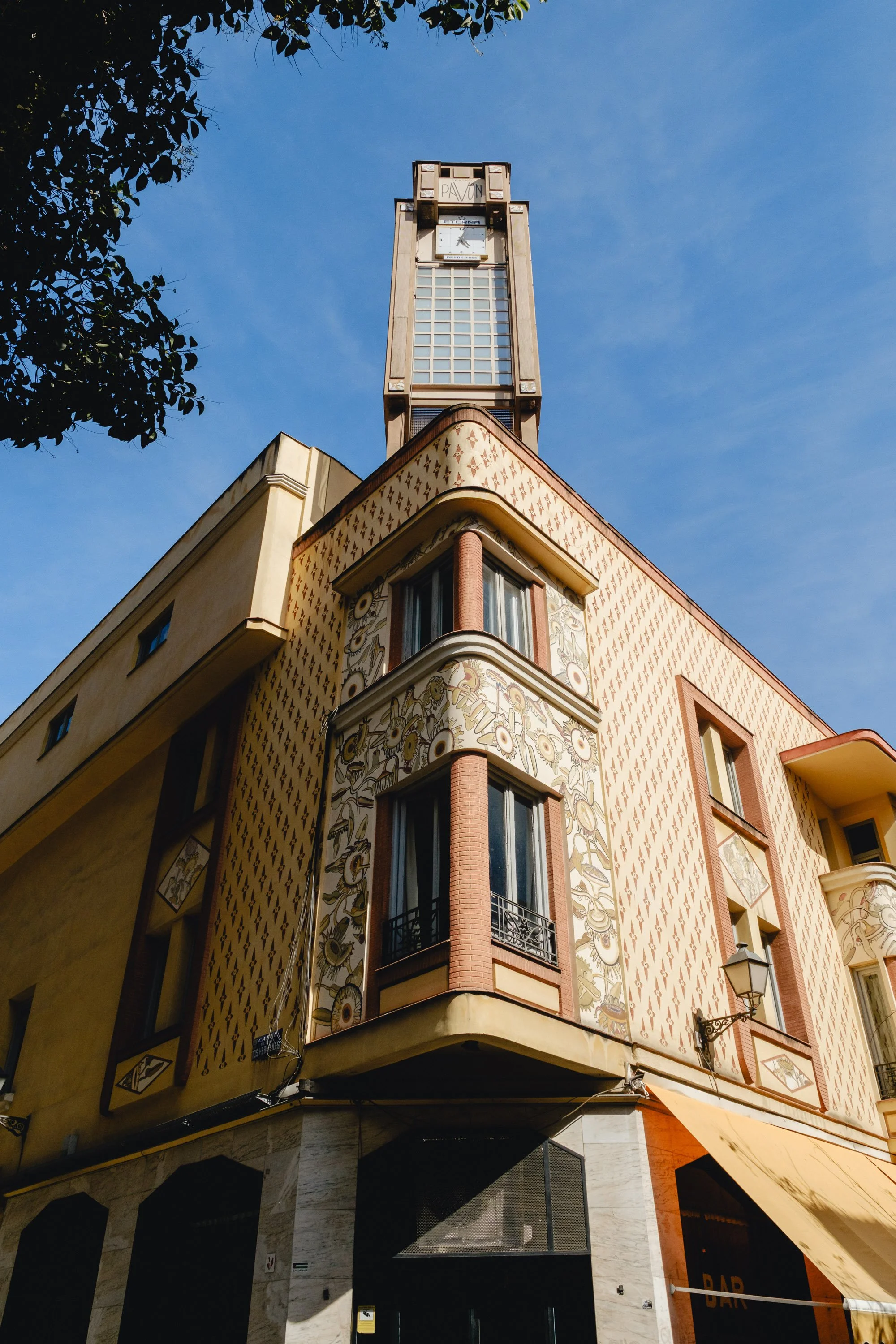 A building with artistic exterior featuring floral and geometric designs, large windows with rounded corners, and a prominent clock tower at the top, set against a blue sky.