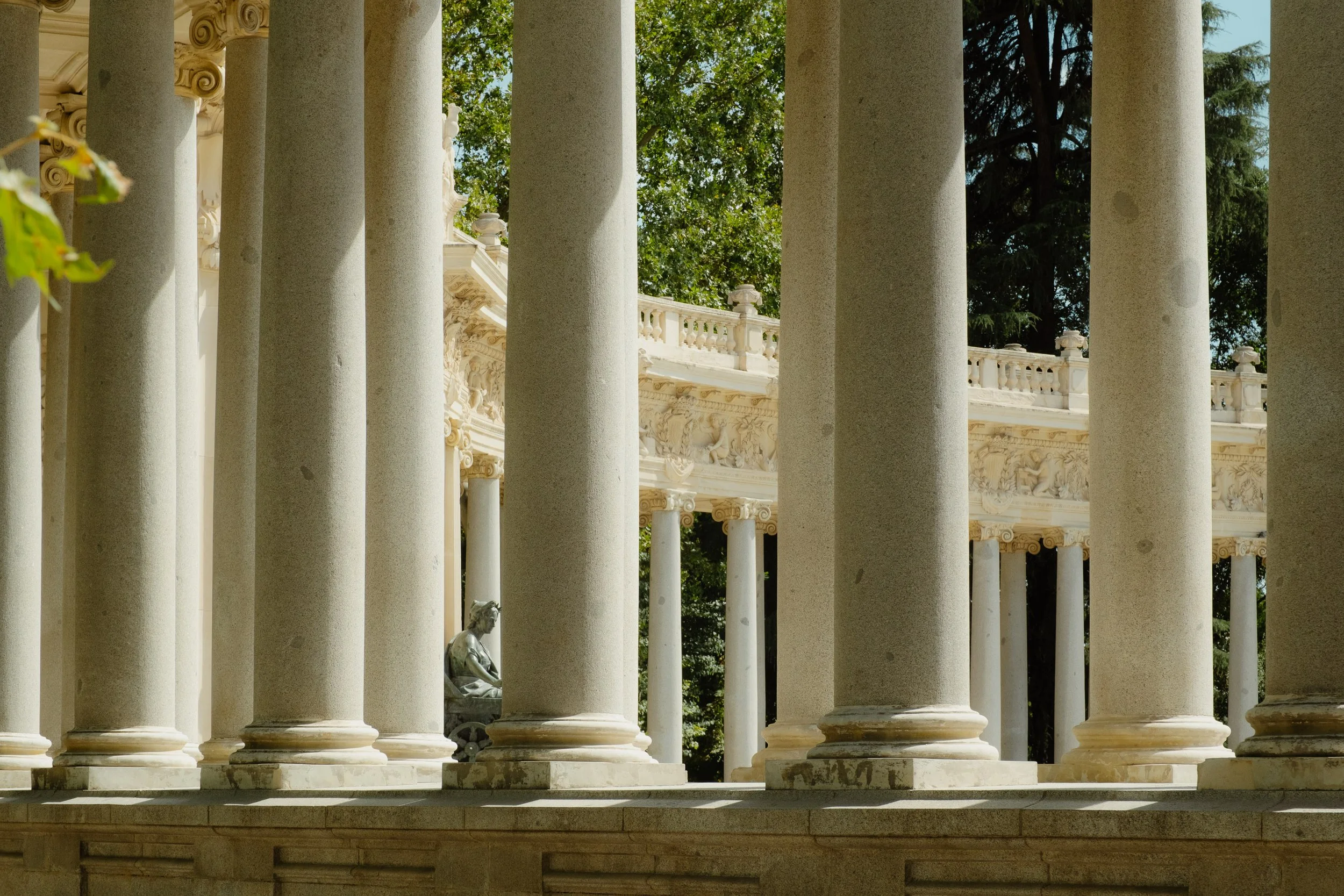 A row of classical marble columns with ornate capitals, part of a Greek or Roman-style structure, with a decorative frieze and trees in the background.