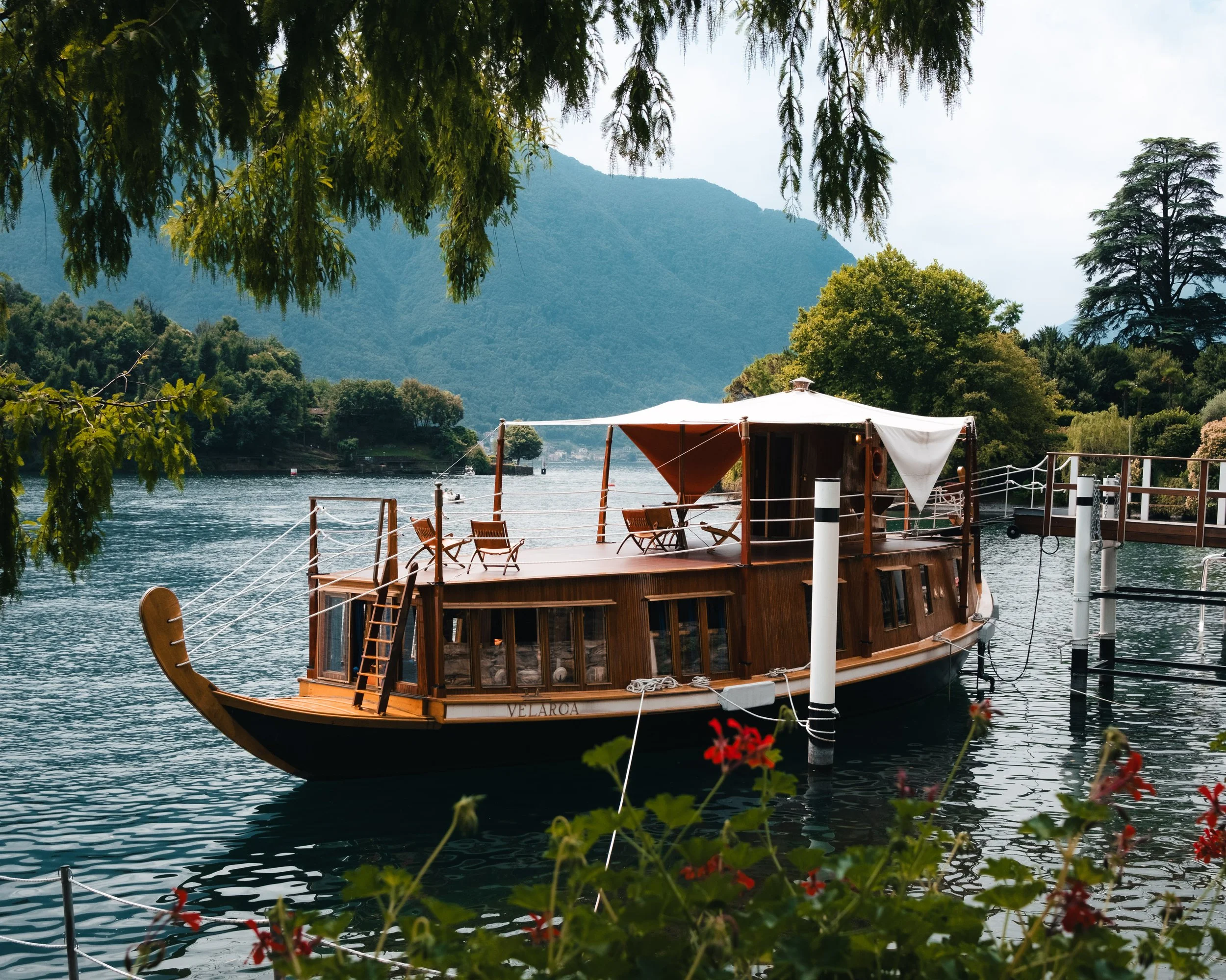 A wooden boat docked on a calm river with lush green trees and hills in the background, and flowers in the foreground.