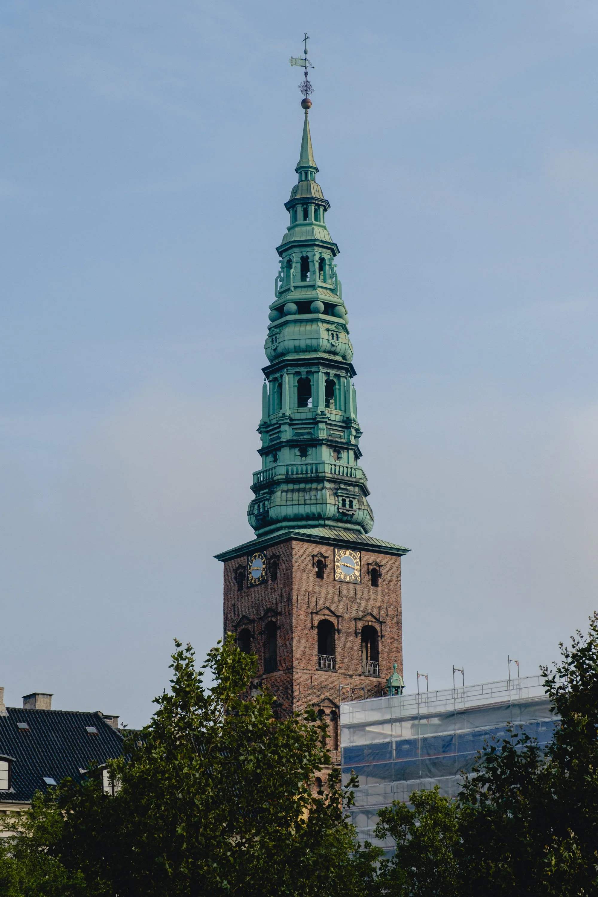 Tall church steeple with a clock and weather vane on top, partly obscured by trees.