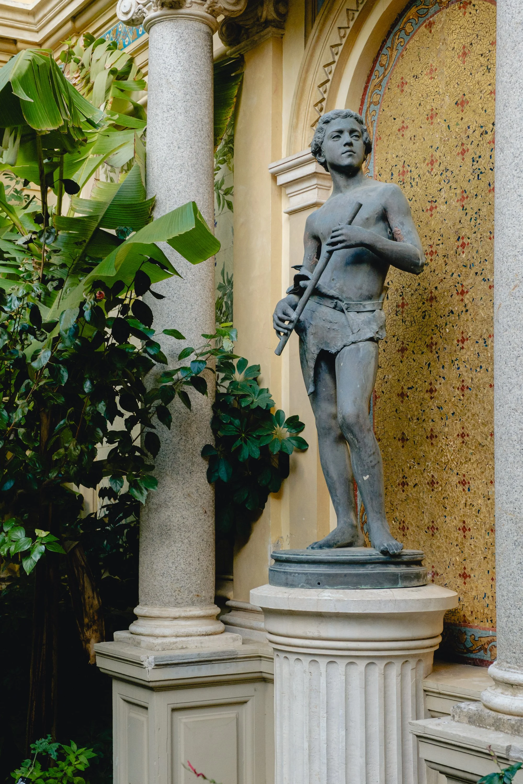 A stone statue of a young boy holding a staff, standing on a pedestal in a lush indoor garden with columns and decorative mosaic background.