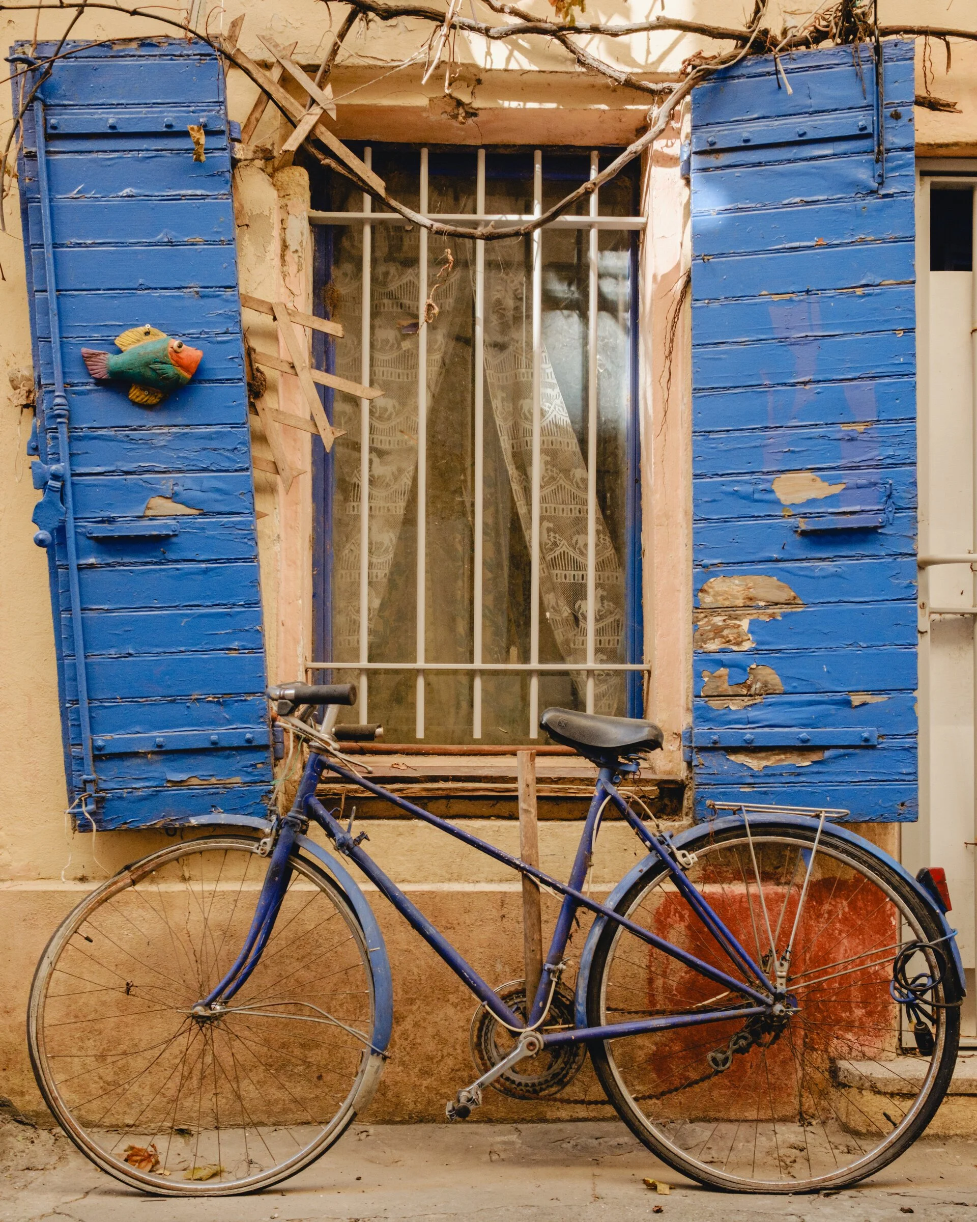 A weathered bicycle leans against a building with peeling blue shutters and a painted fish ornament in Arles