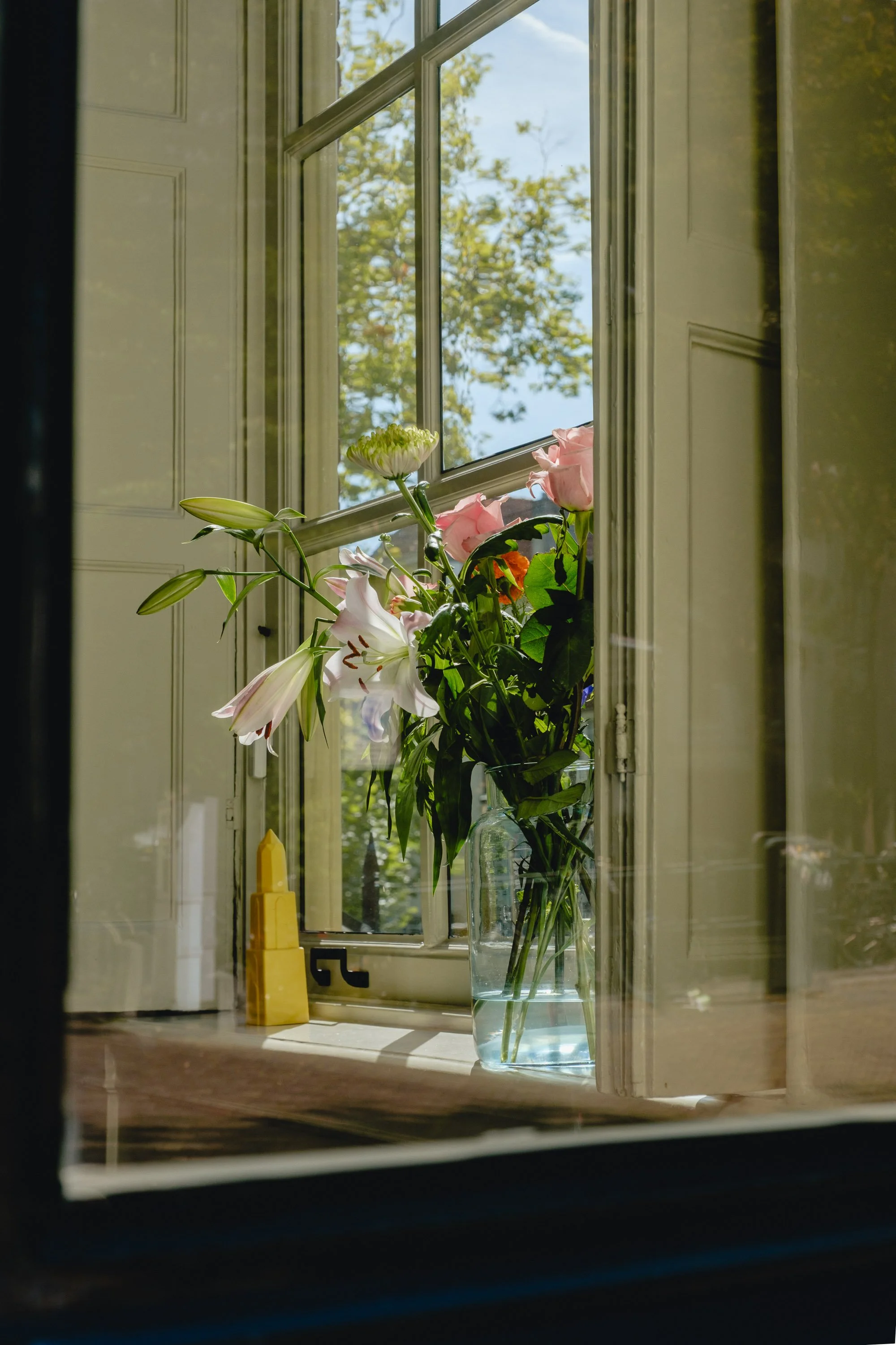 A window on a wall with a vase of assorted flowers on the windowsill, sunlight streaming in, and a tree with green leaves visible outside.
