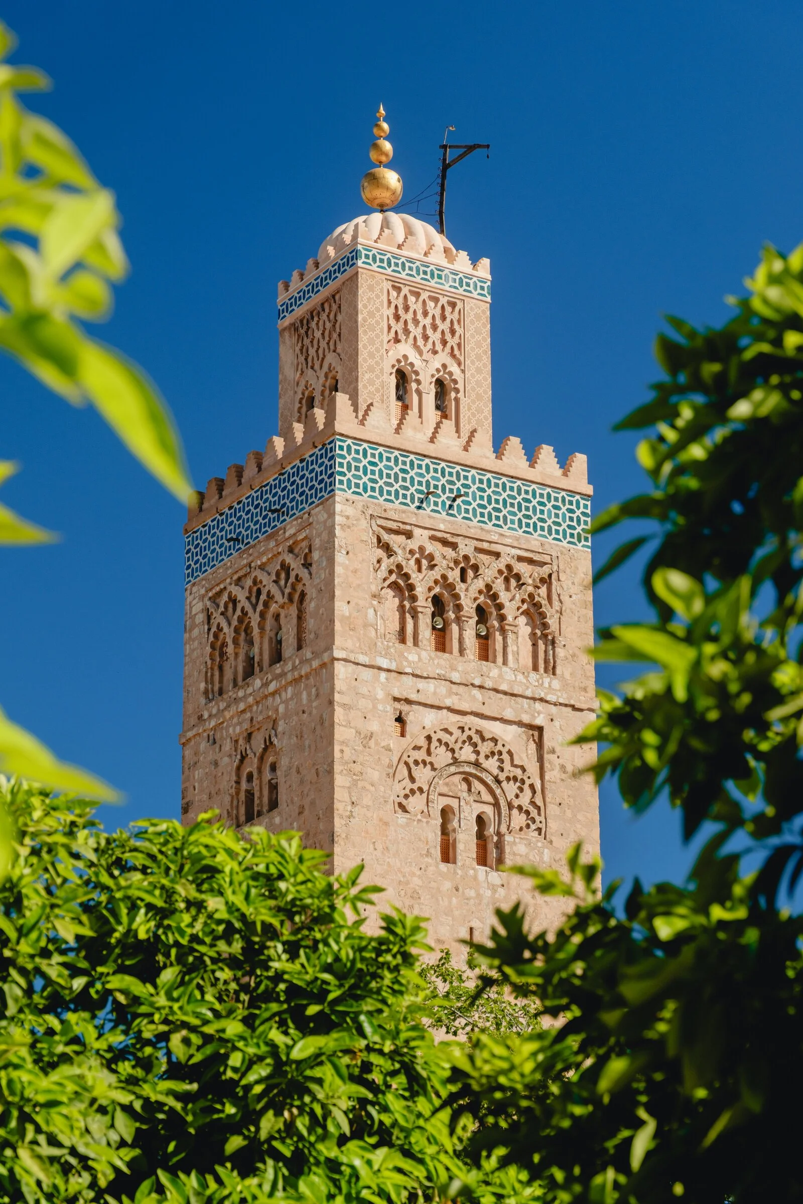 The Koutoubia Mosque's ornate minaret rises above green treetops against a deep blue sky