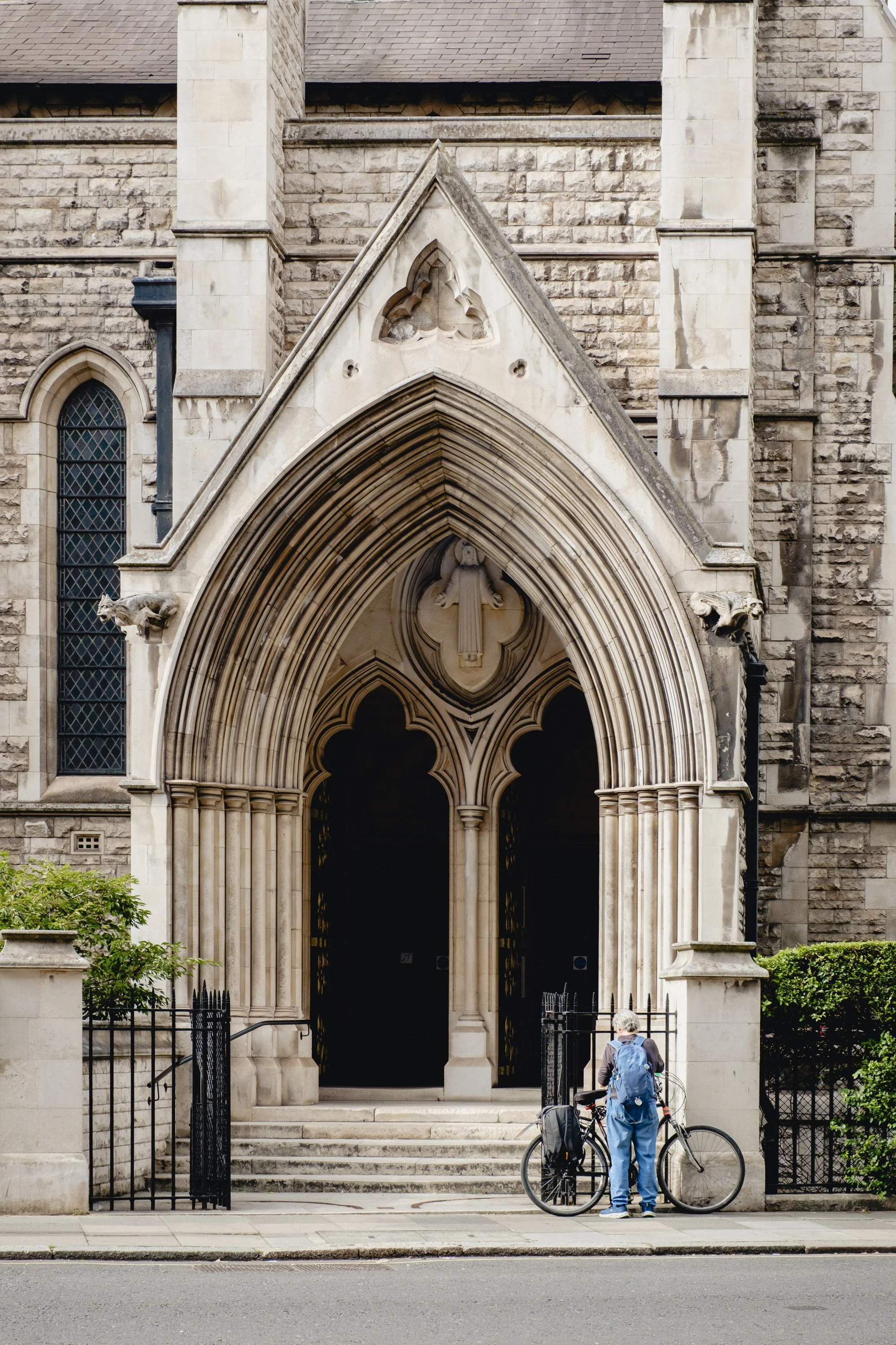 A person standing with a bicycle in front of a gothic church entrance with pointed arch, stone columns, and decorative sculptures.