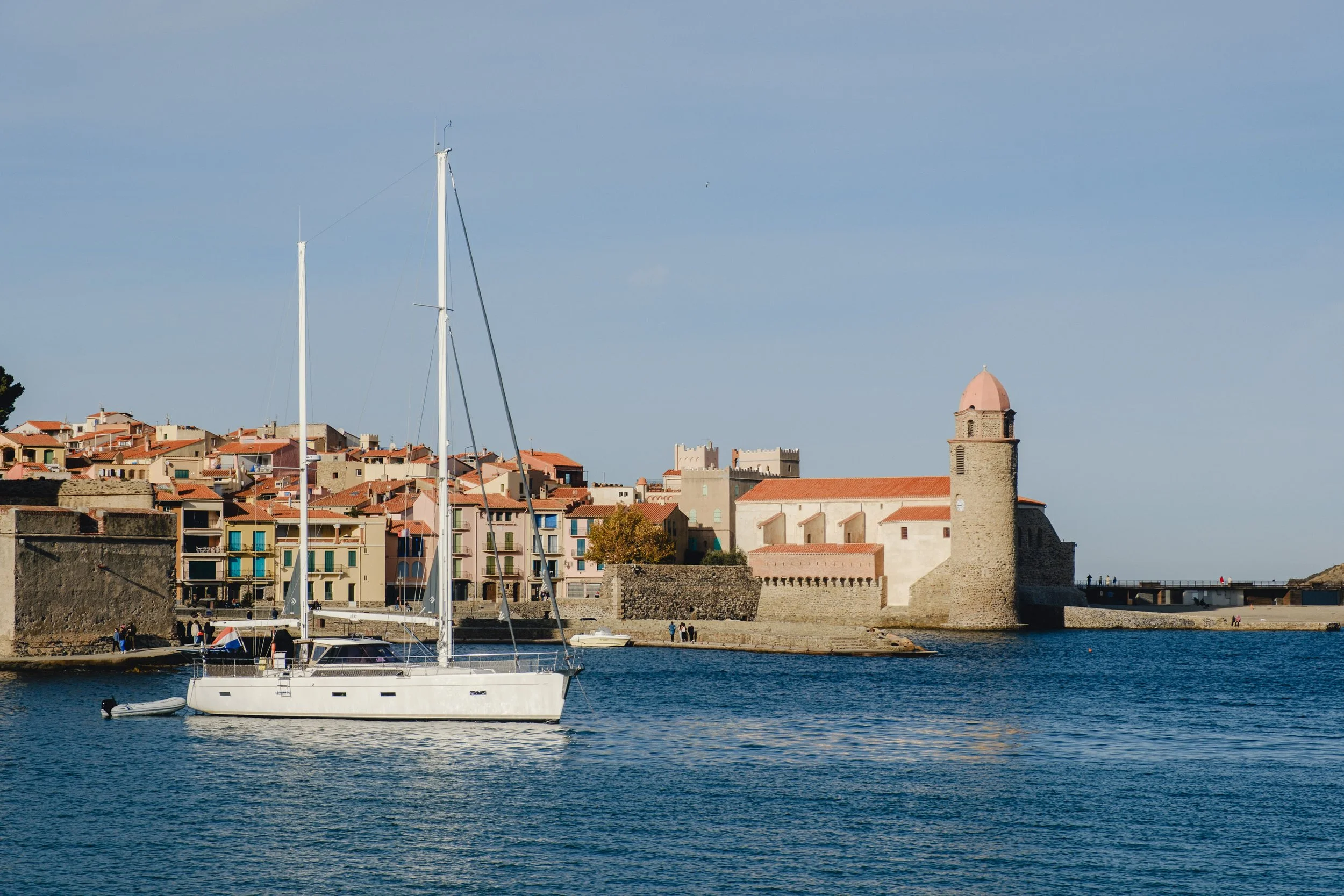 A sailboat floating on the water near a historic coastal town with colorful buildings and a stone castle wall.