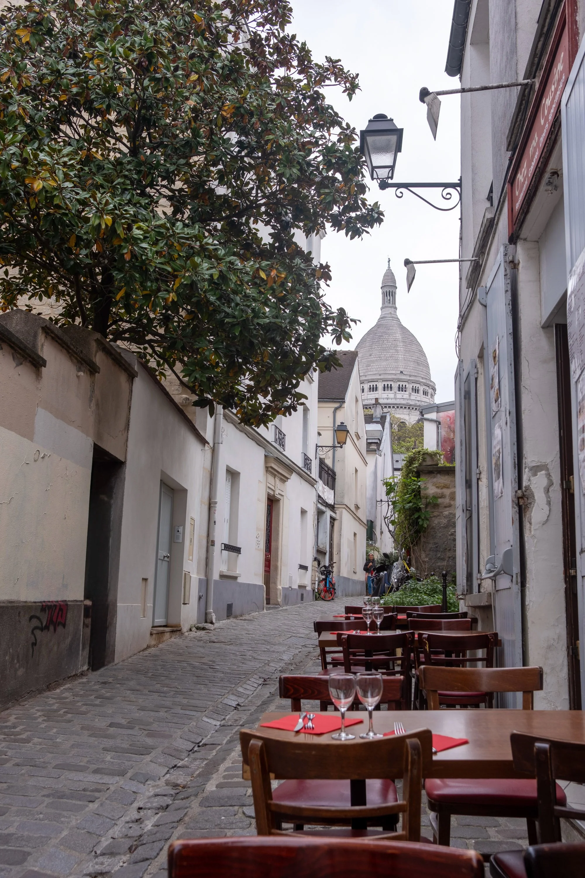A cobblestone street with outdoor restaurant seating, featuring wooden tables and chairs with wine glasses and red napkins, with the Sacré-Cœur Basilica visible in the background in Paris.