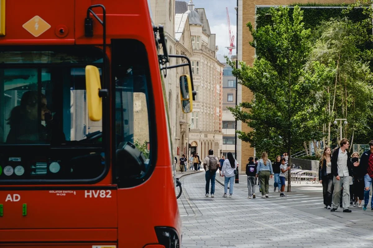 A red London bus rounds a corner near a modern pedestrian square