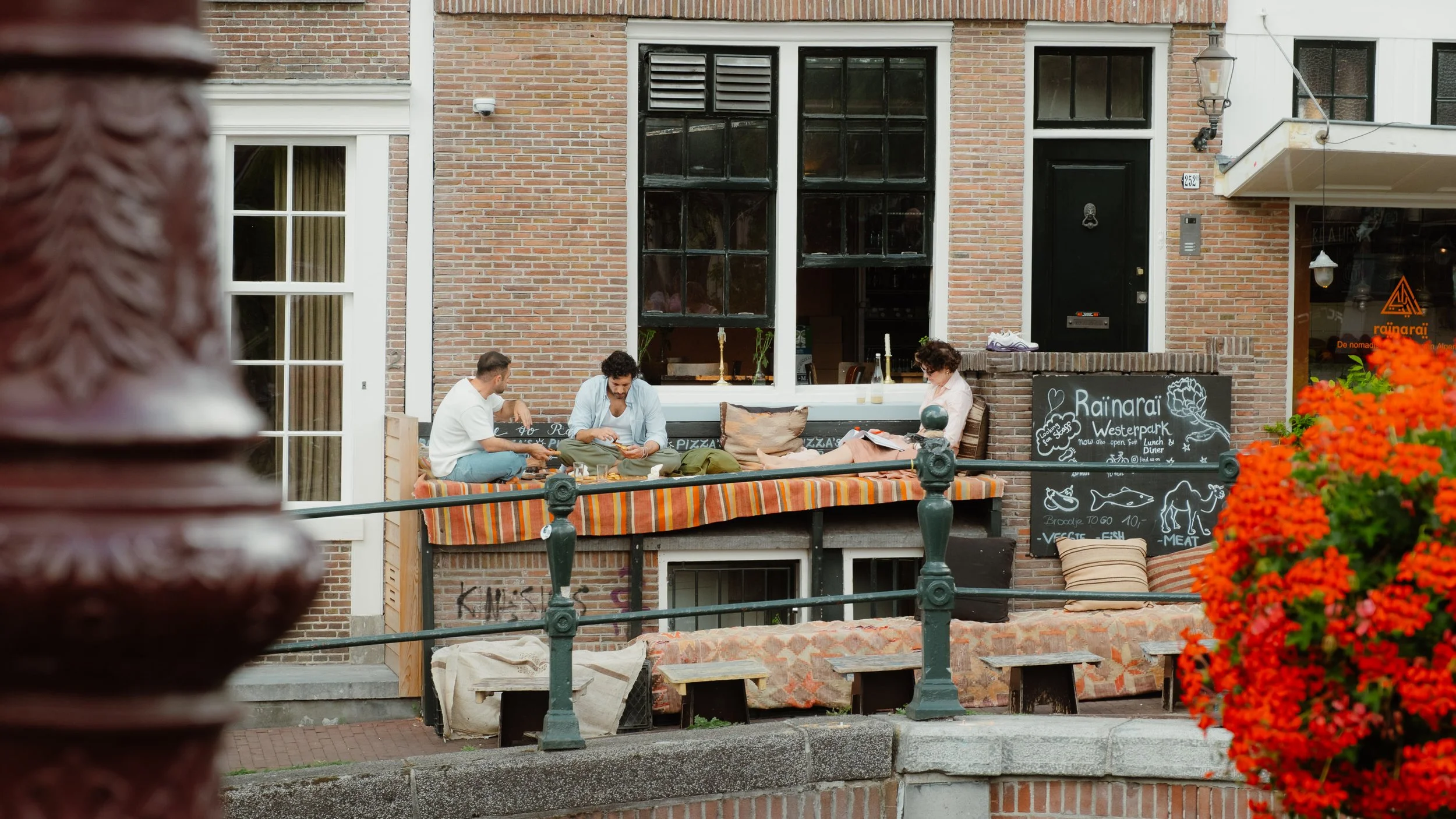 Three people sitting on a decorated outdoor terrace of a cafe or restaurant, with a brick building in the background, black framing around the windows, and a black chalkboard sign with menu drawings and text.
