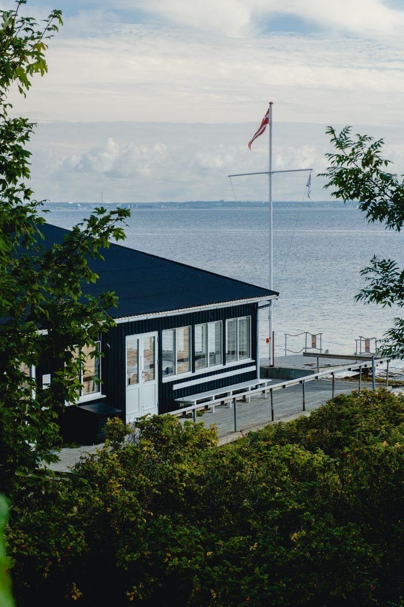 A dark wooden boathouse on a pier flying a Danish flag, overlooking the open sea