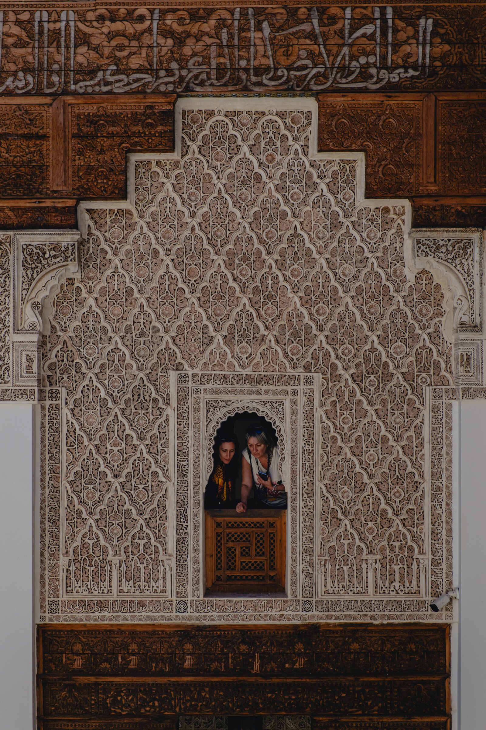 Two visitors lean out from an ornately carved stucco archway window in a Marrakech madrasa