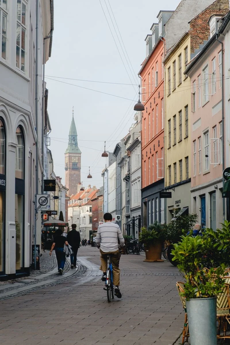 A cyclist rides down a cobbled shopping street toward a distant church spire, flanked by colorful facades