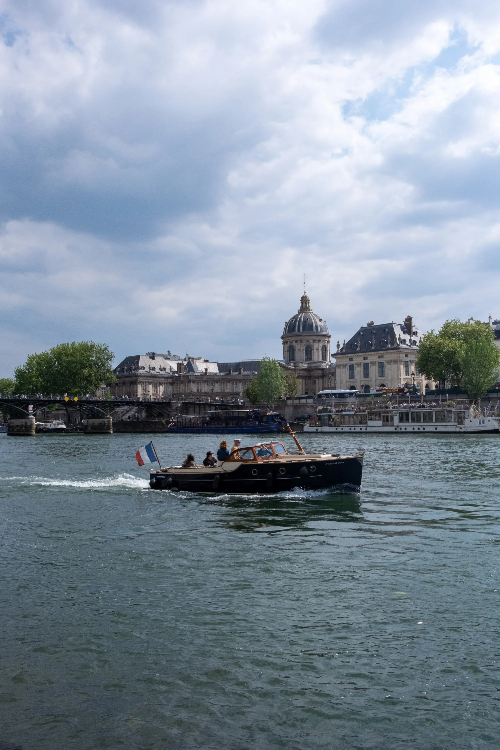 A boat sailing on a river with historic buildings and a domed church in the background, under a partly cloudy sky.