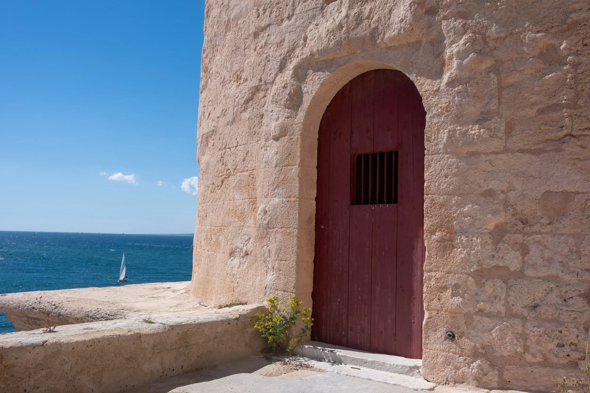 A stone fortress wall with a red wooden door and barred window near the coast. The background shows the ocean with a sailboat and a partly cloudy sky.