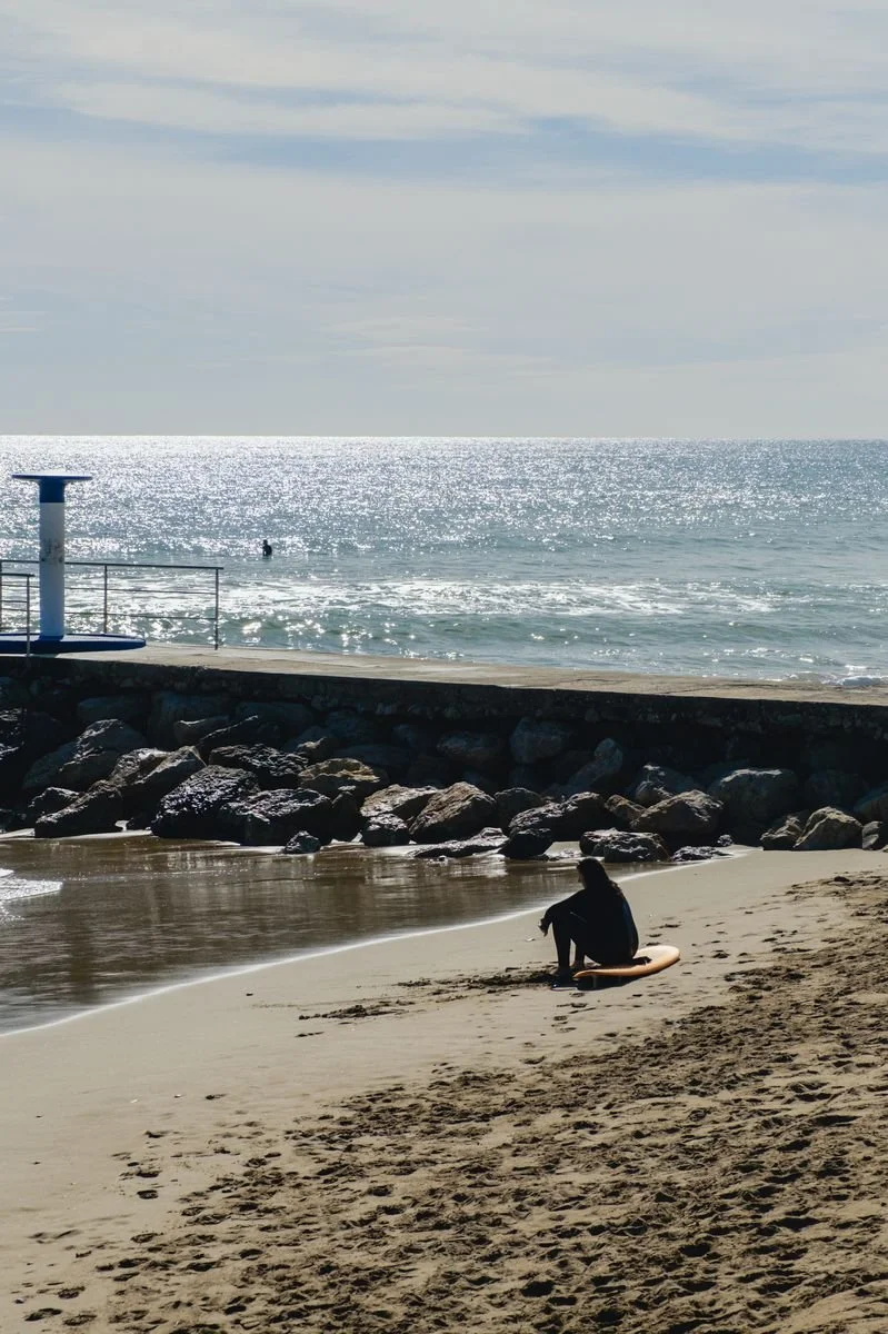 A surfer sits on their board at the shoreline, the sea glittering behind
