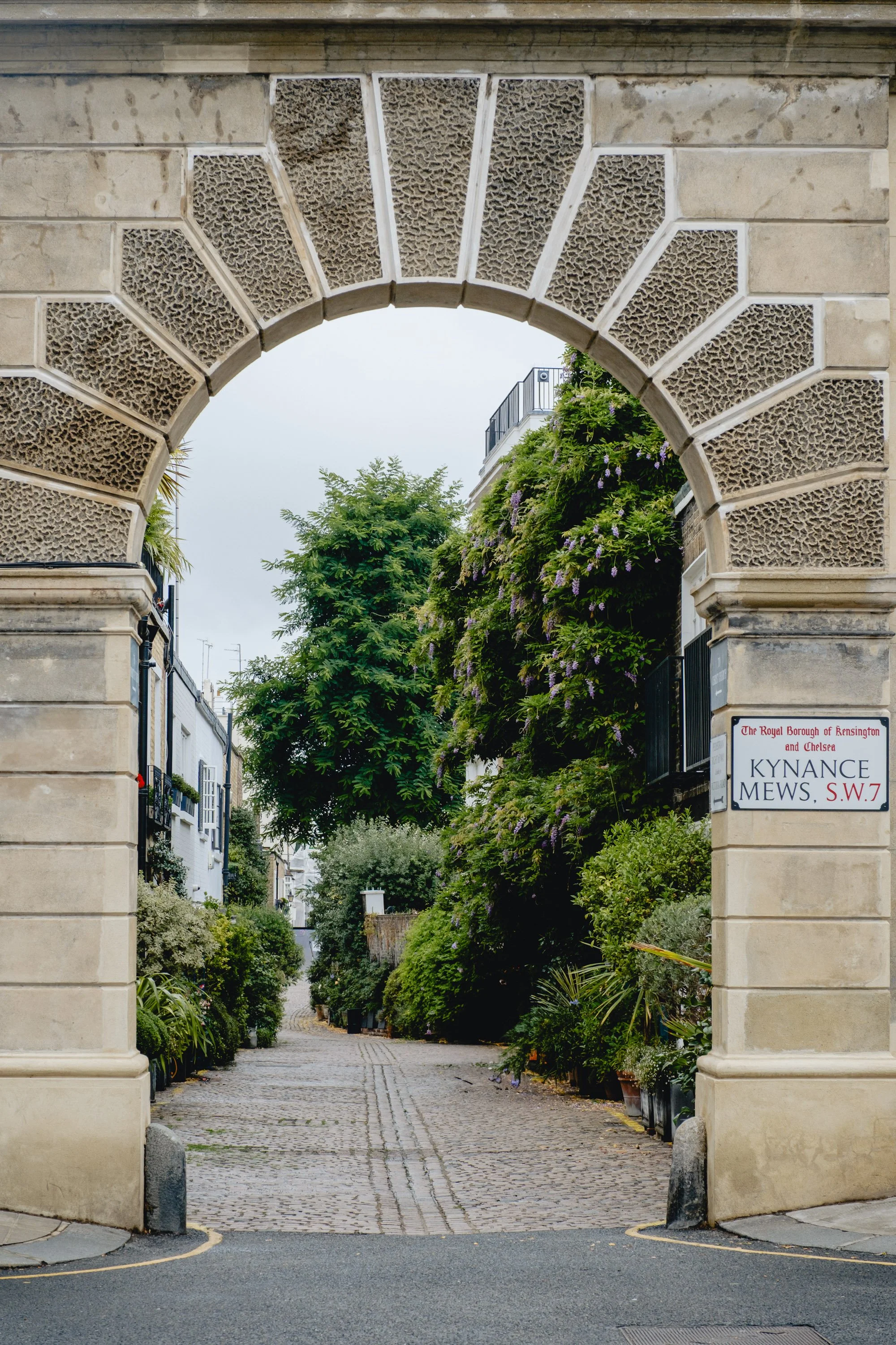 A stone archway leading into a cobblestone alley lined with lush green plants and trees, with a sign indicating the location in Kensington and Chelsea.