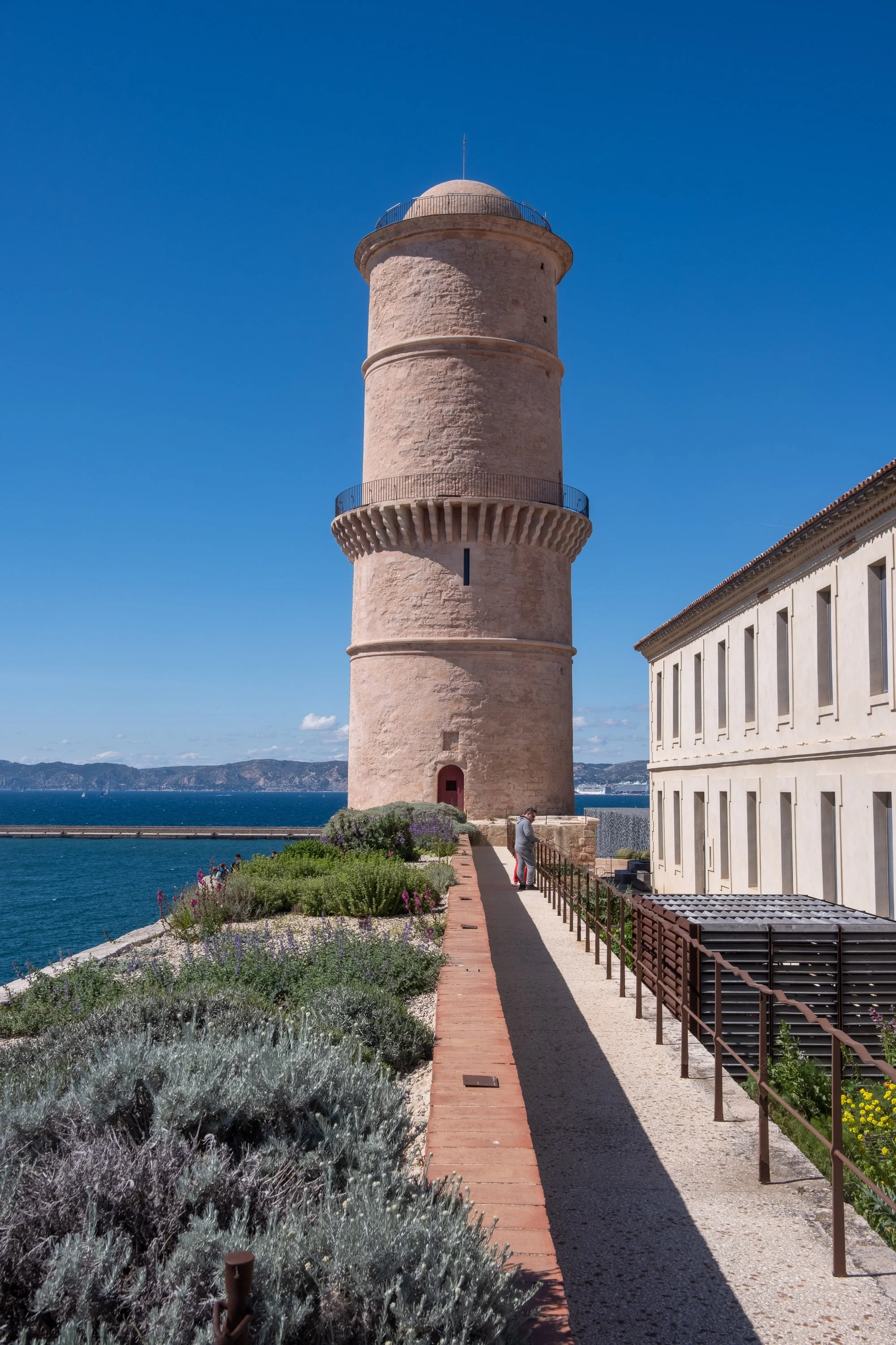 A tall, circular stone lighthouse on the waterfront under a clear blue sky, with some people near its base and a garden with plants and flowers along a path.