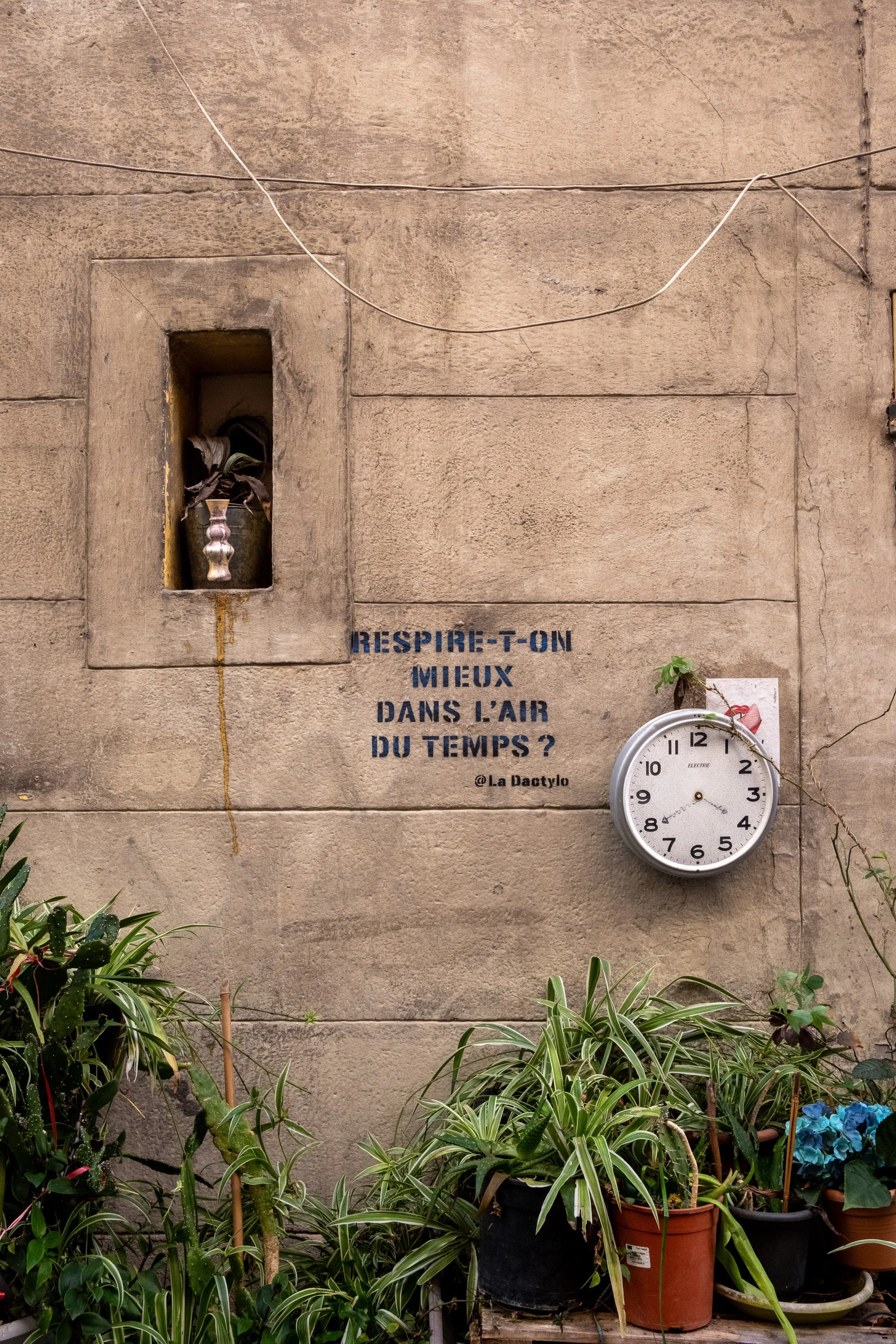 A stone wall with a small niche containing a vase with flowers, a clock hanging next to a poster, plants at the bottom, and a French quote painted on the wall.