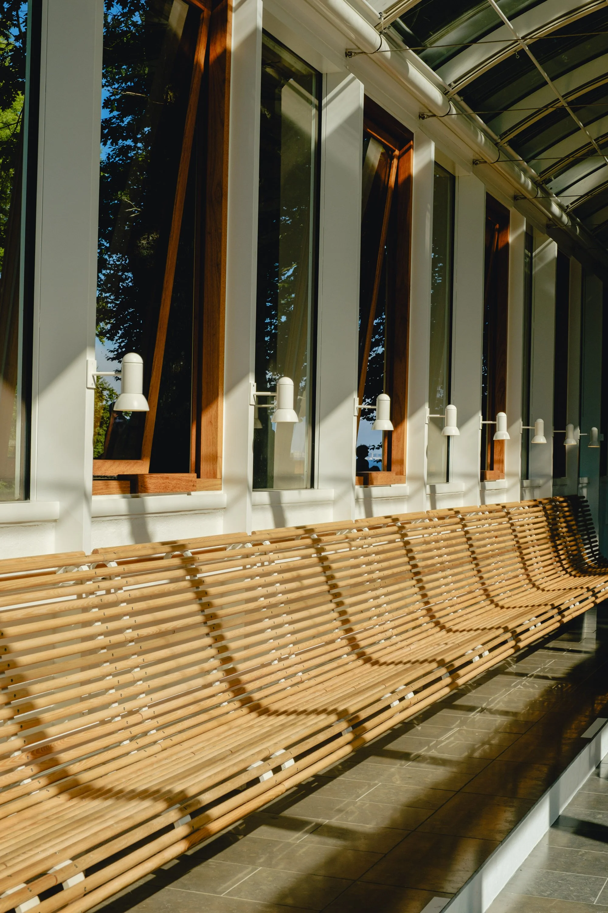 Sunlight filters through a row of arched wooden windows in a modern building, casting shadows on a long wooden bench along the wall.