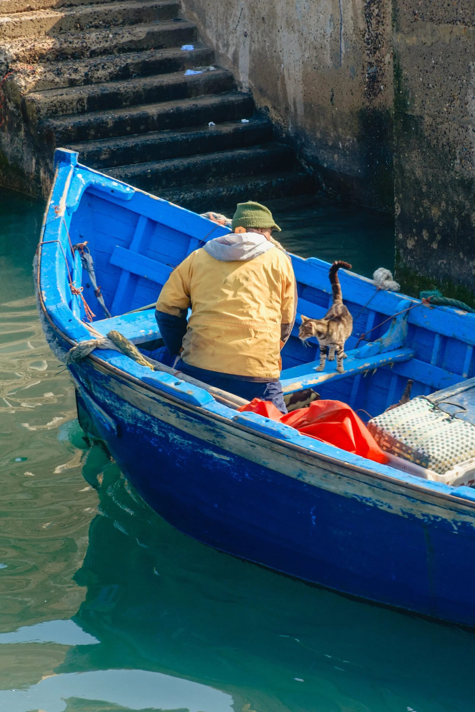 A fisherman in a yellow jacket stands in his blue boat beside his tabby cat