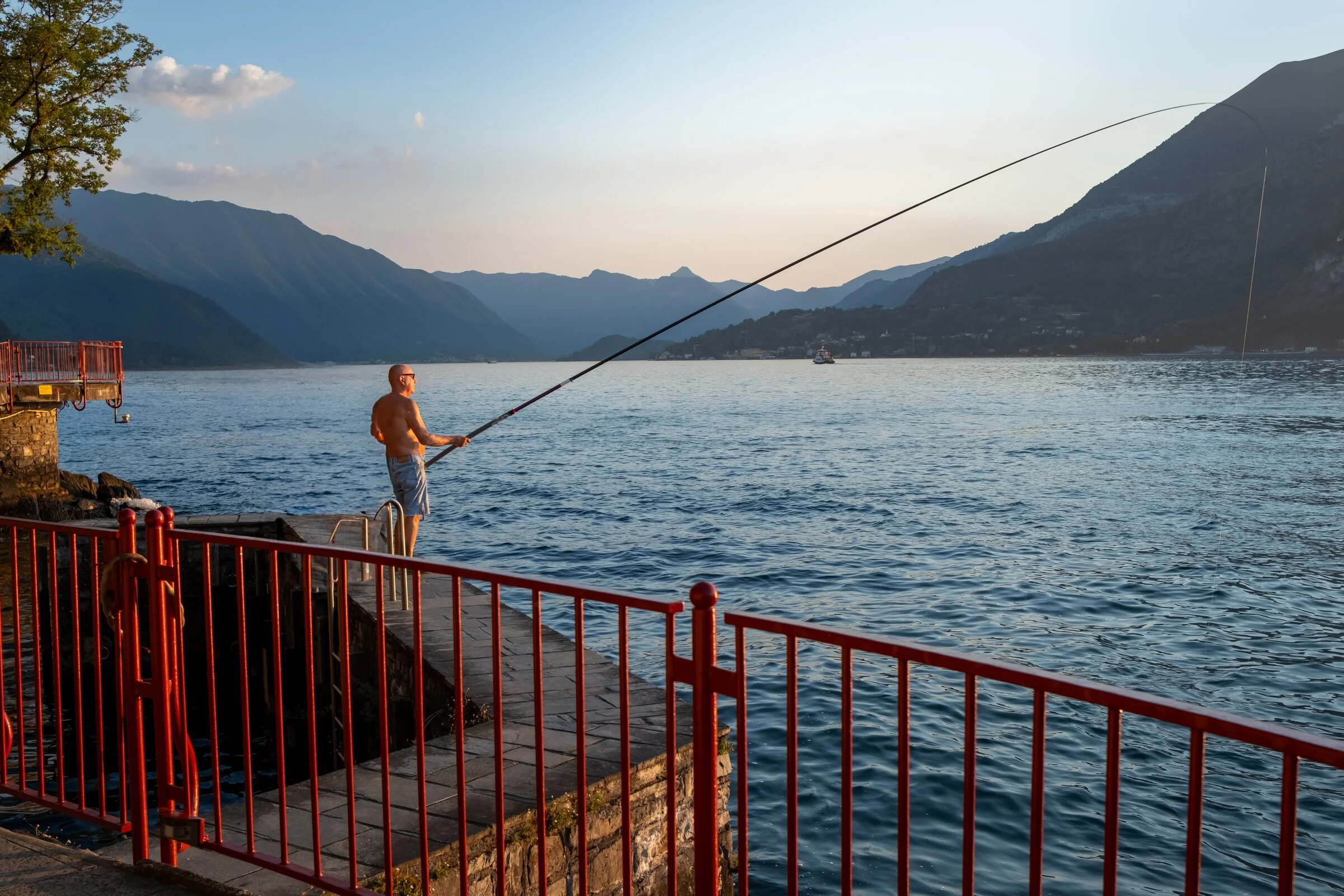 A shirtless man casts a fishing line from a red-railed pier into Lake Como as the sun sets over the mountains
