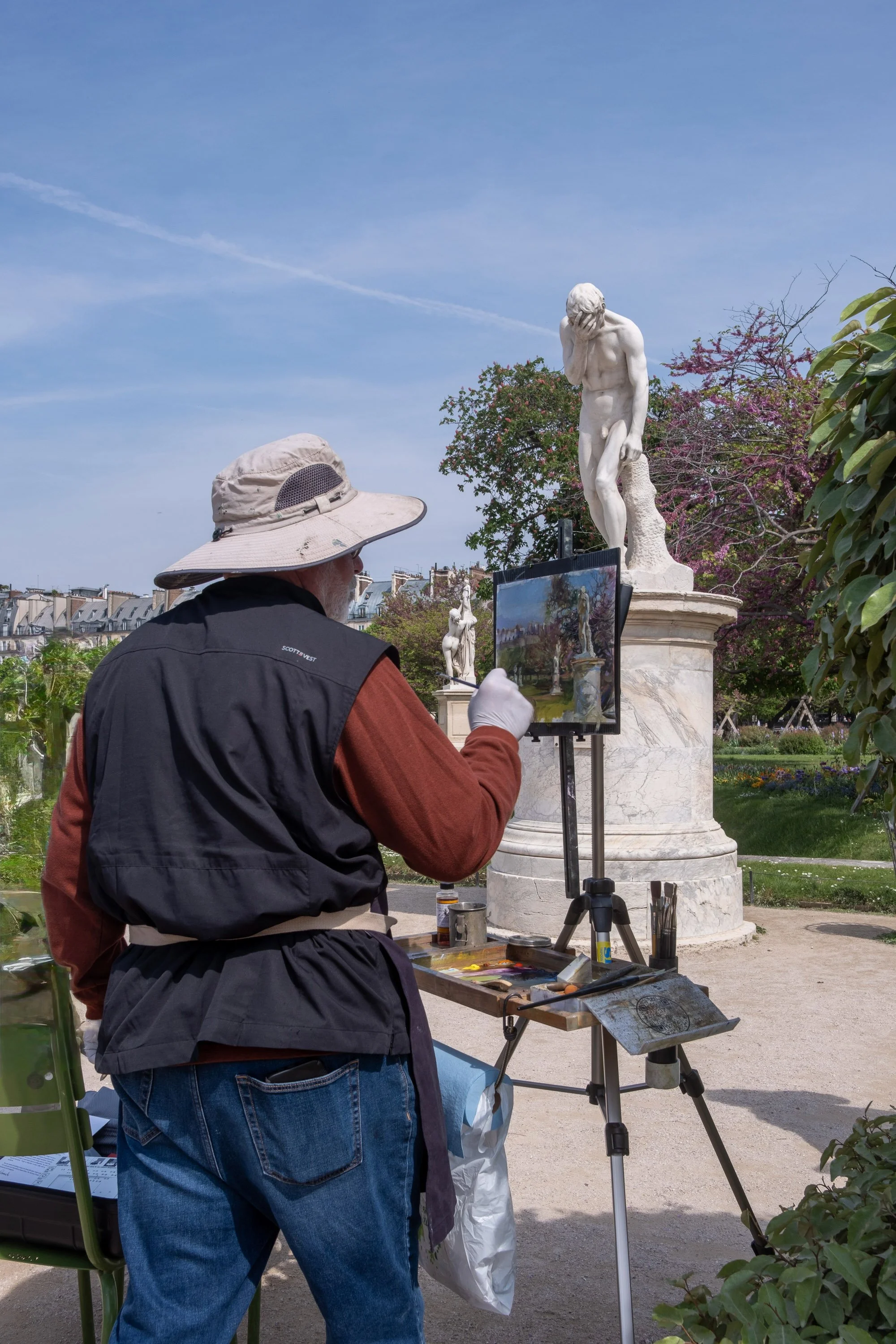 An artist painting in a park with classical sculptures and blooming trees in the background.