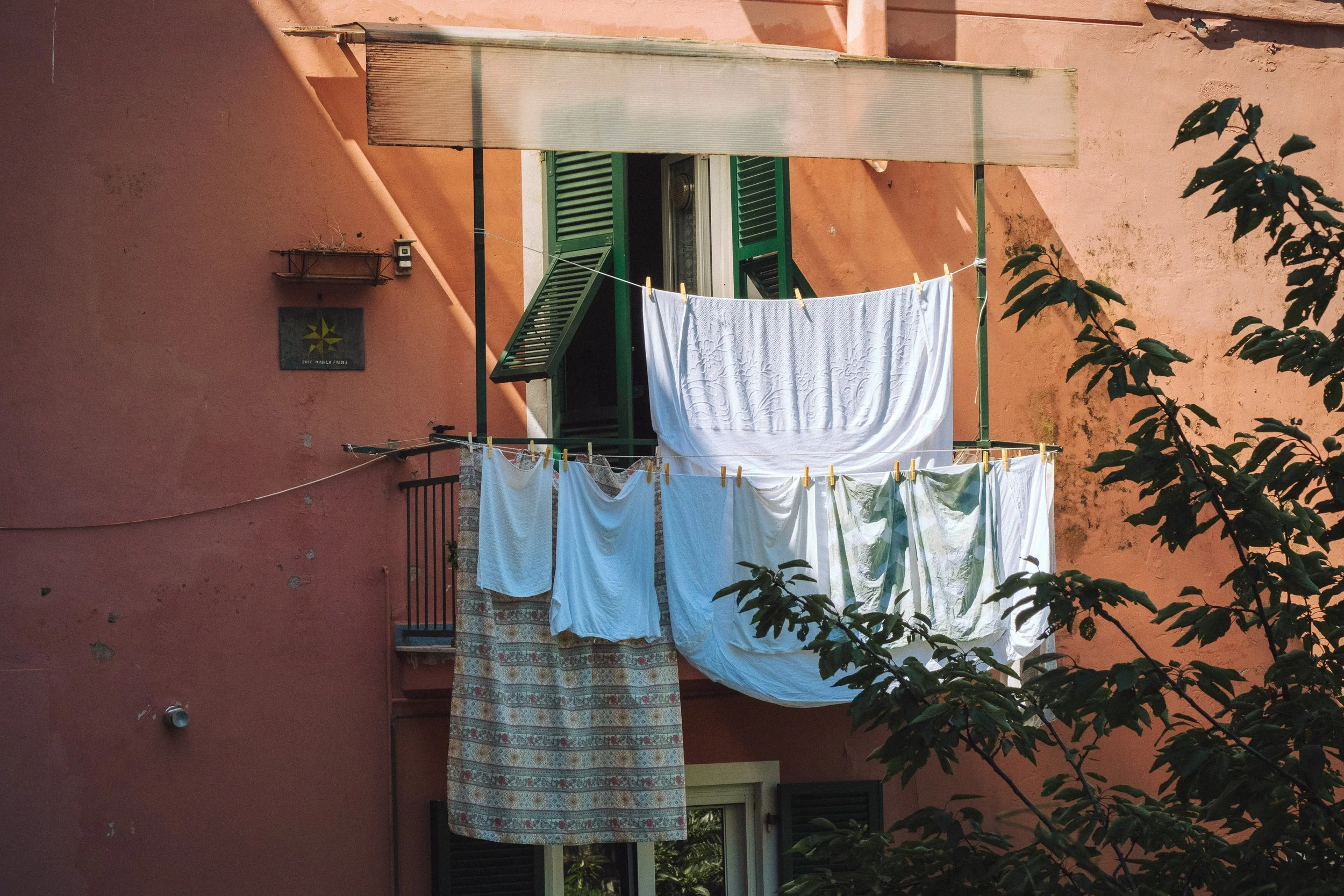 Laundry hanging on a clothesline outside a pink building with green shutters, sunlight casting shadows.