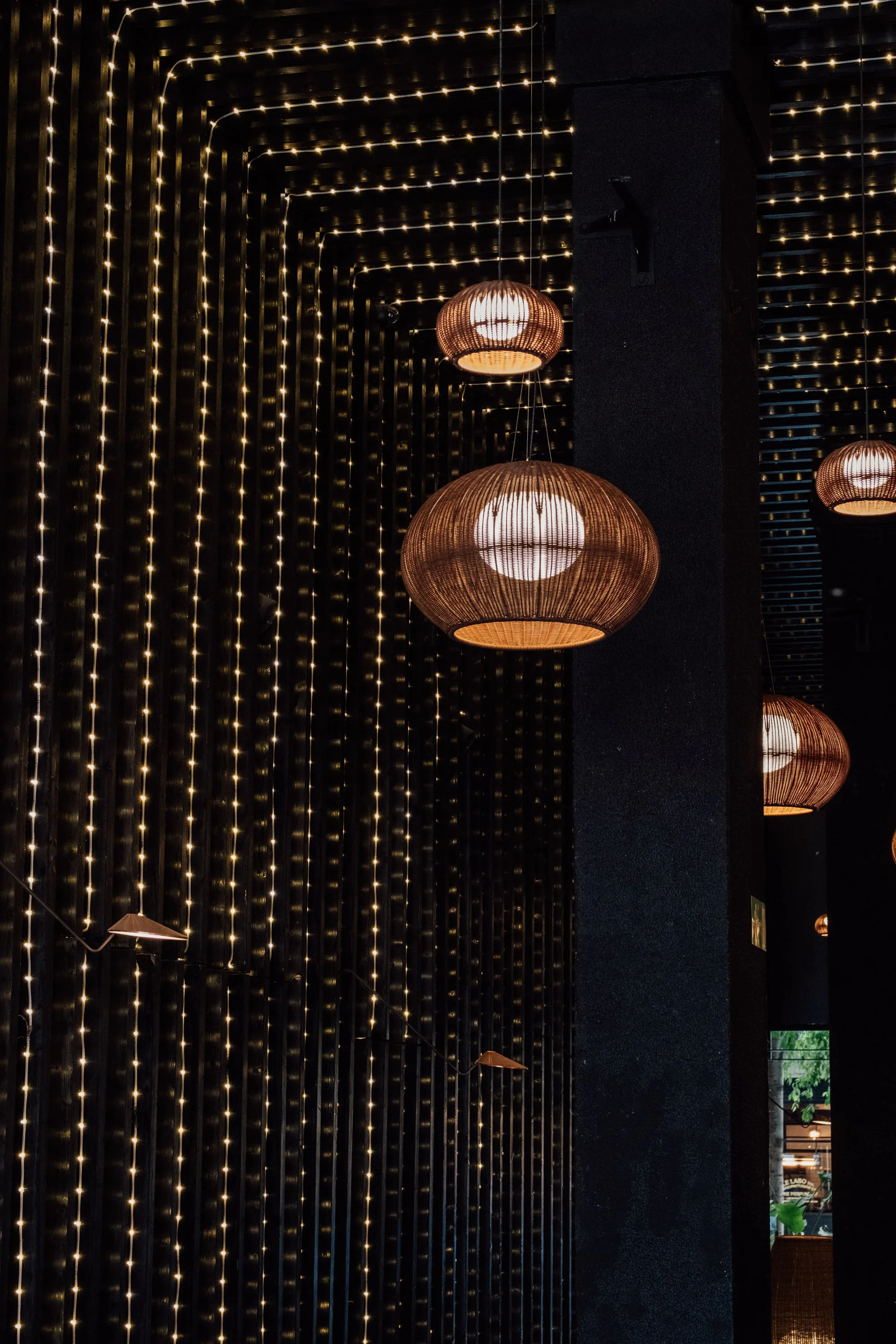 Interior of a dimly lit space with hanging woven pendant lights and string lights on a black wall.