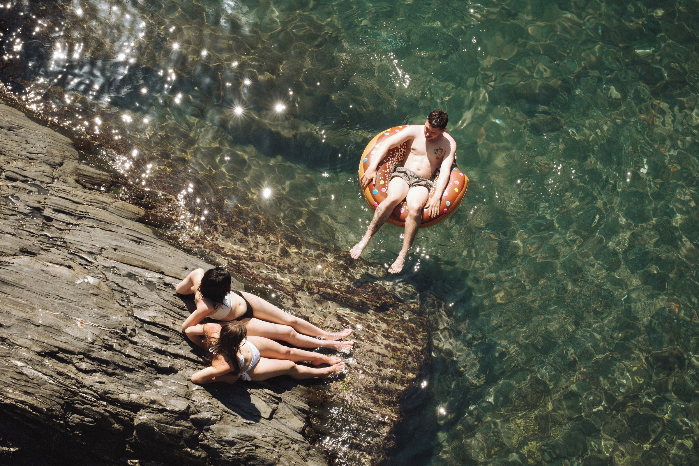 A family lounges on rocks while a man floats in an inflatable ring in clear green water, seen from above