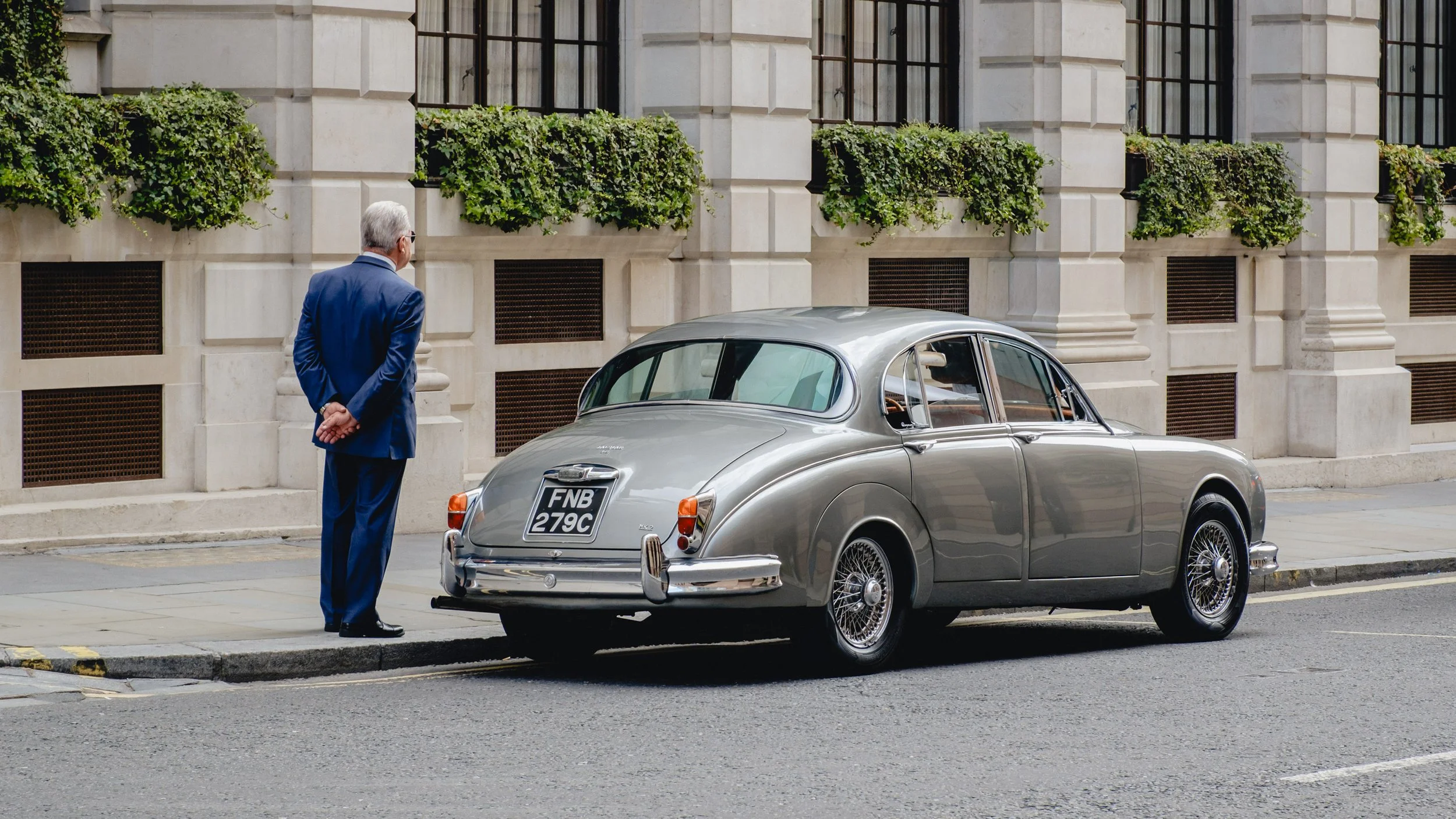 A man in a blue suit is standing on the sidewalk looking at a vintage silver car parked by the curb in front of a building with large windows and green plants.