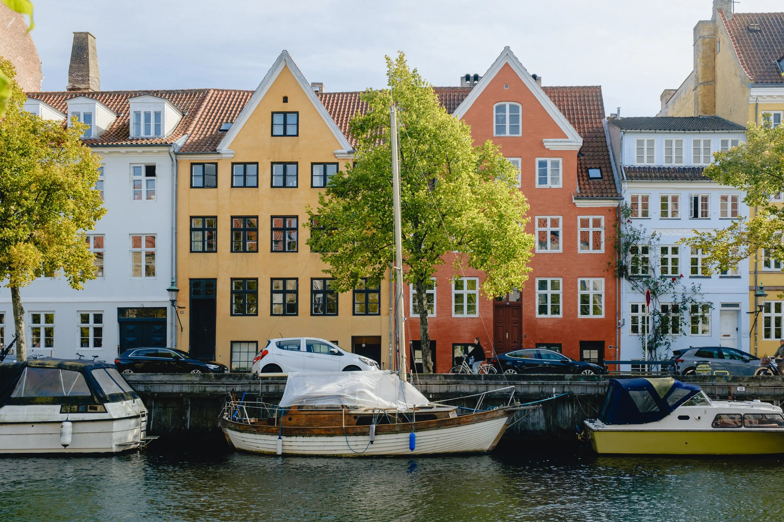 Colorful row of European-style buildings with trees and parked cars along a canal, boats docked at the water's edge.