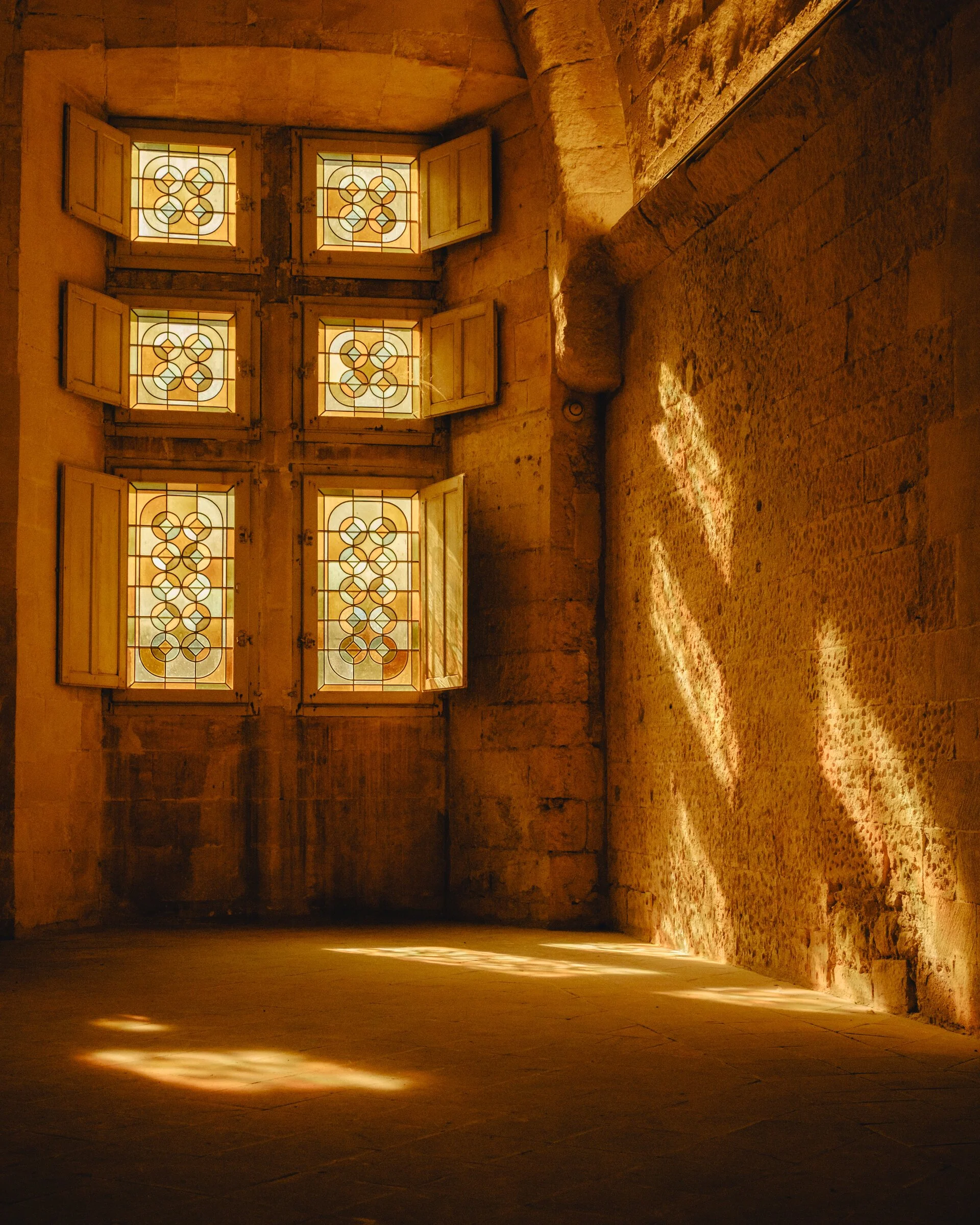 Warm afternoon light streams through leaded stained-glass windows onto a stone wall