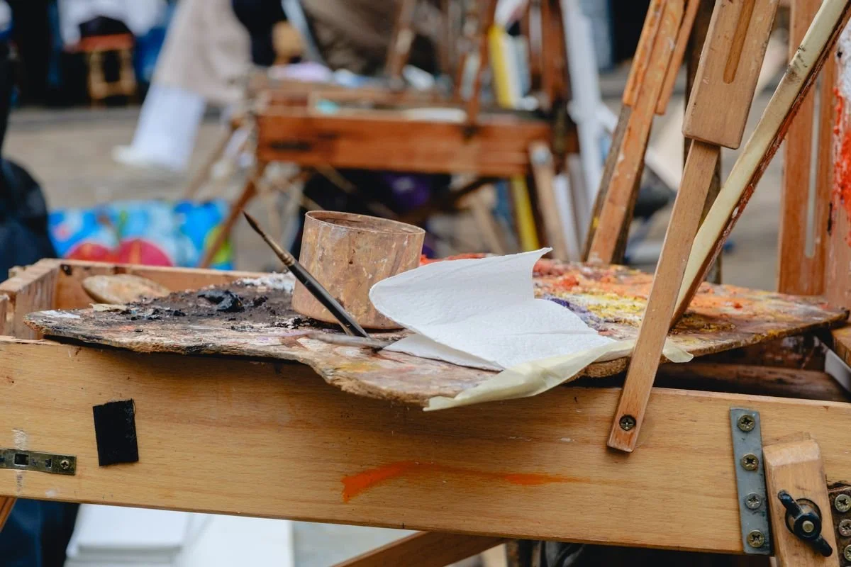 Close-up of a paint-splattered artist's palette, brush, and rag on a wooden easel tray