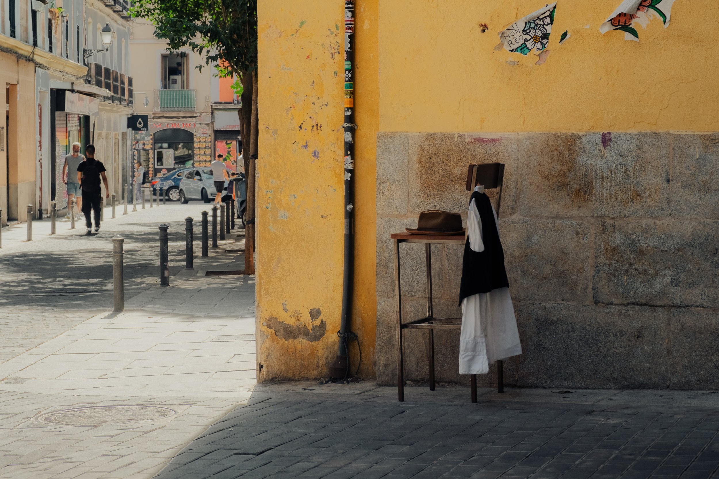 Street scene with a yellow wall, a table with clothing and a hat, pedestrians walking along the sidewalk, parked cars, and colorful buildings in the background.