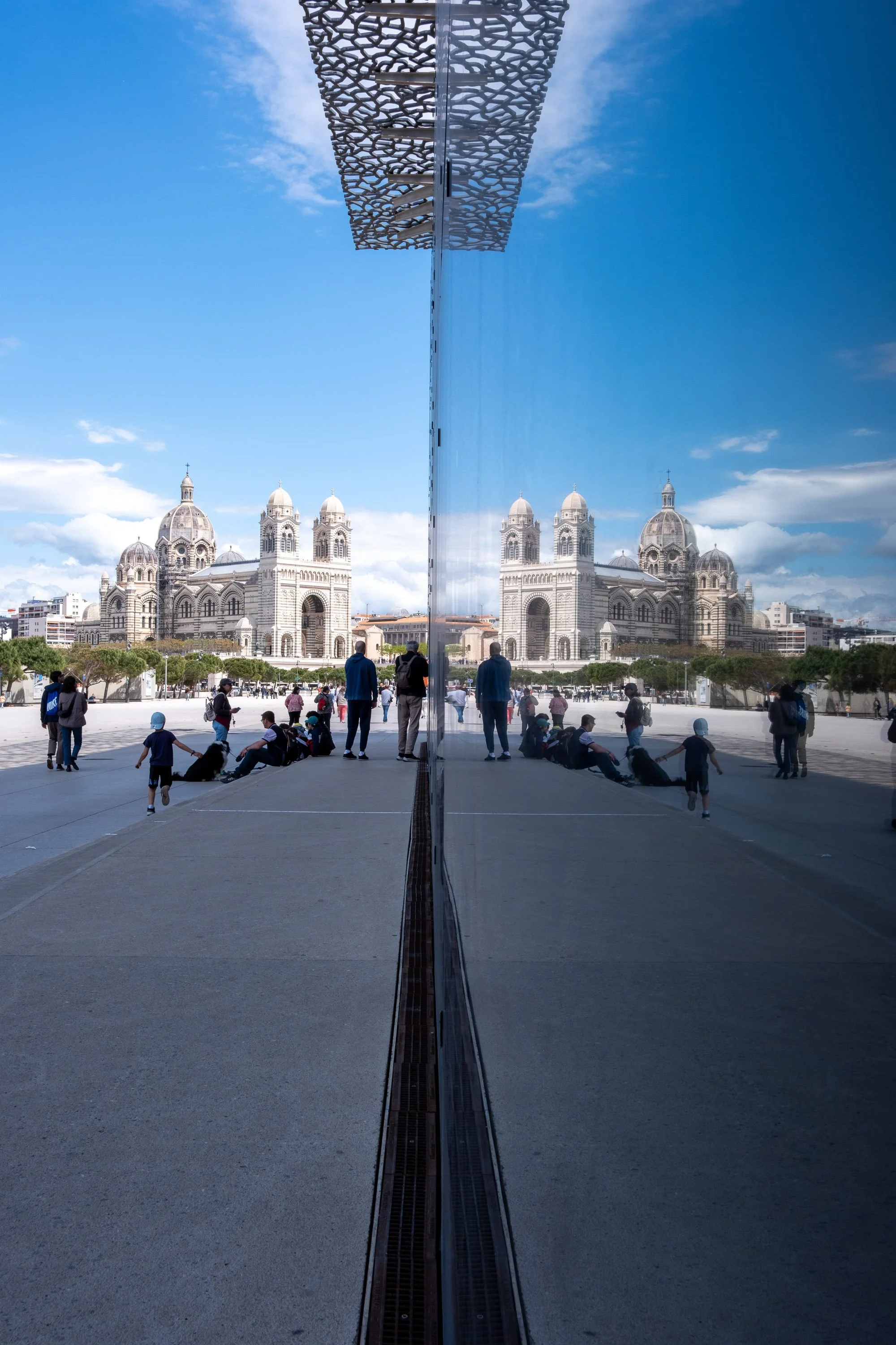 A city square with a large church in the background reflected in a tall, mirror-like building. People are sitting and walking on the square, and the sky is partly cloudy.