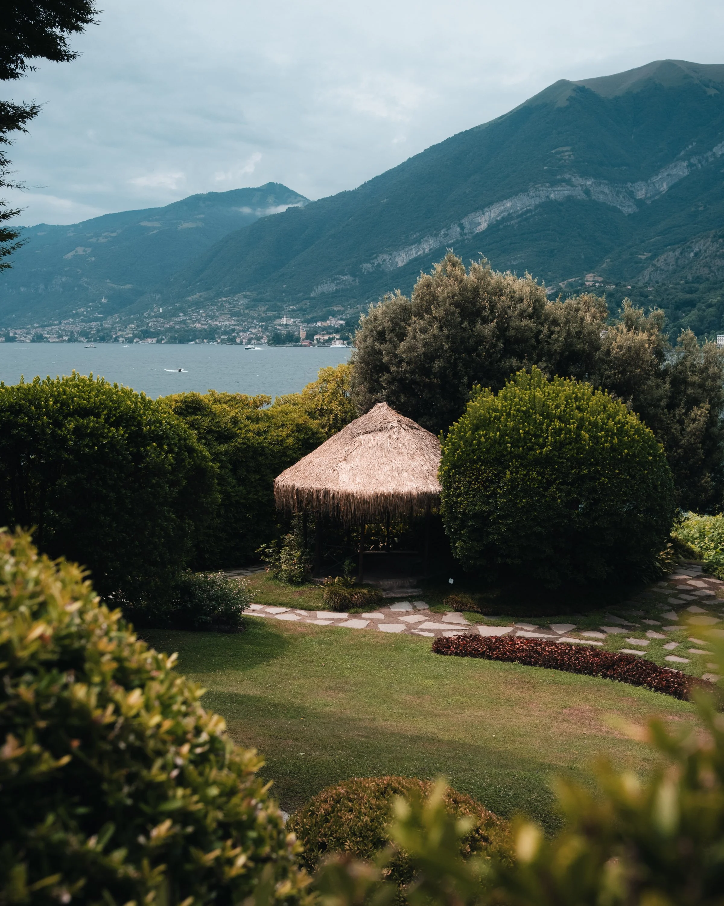 A lakeside scene with green trees, bushes, and a thatched-roof gazebo in the foreground, mountains across the lake, and boats on the water.