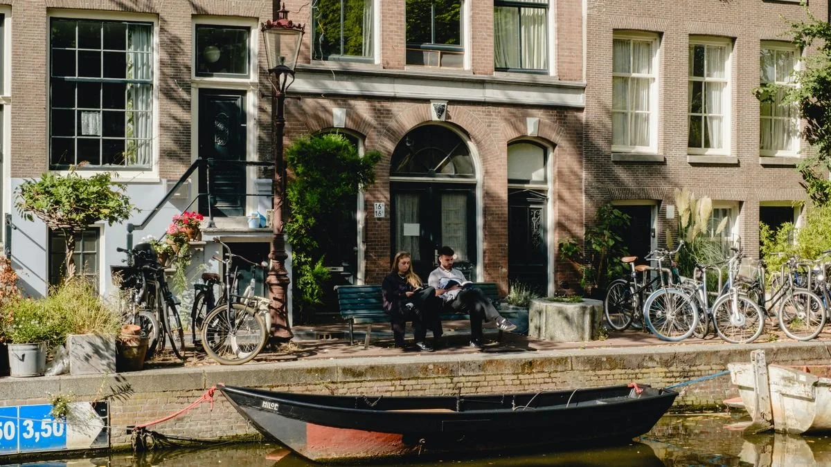 A couple reads together on a canal-side bench, small boats moored below