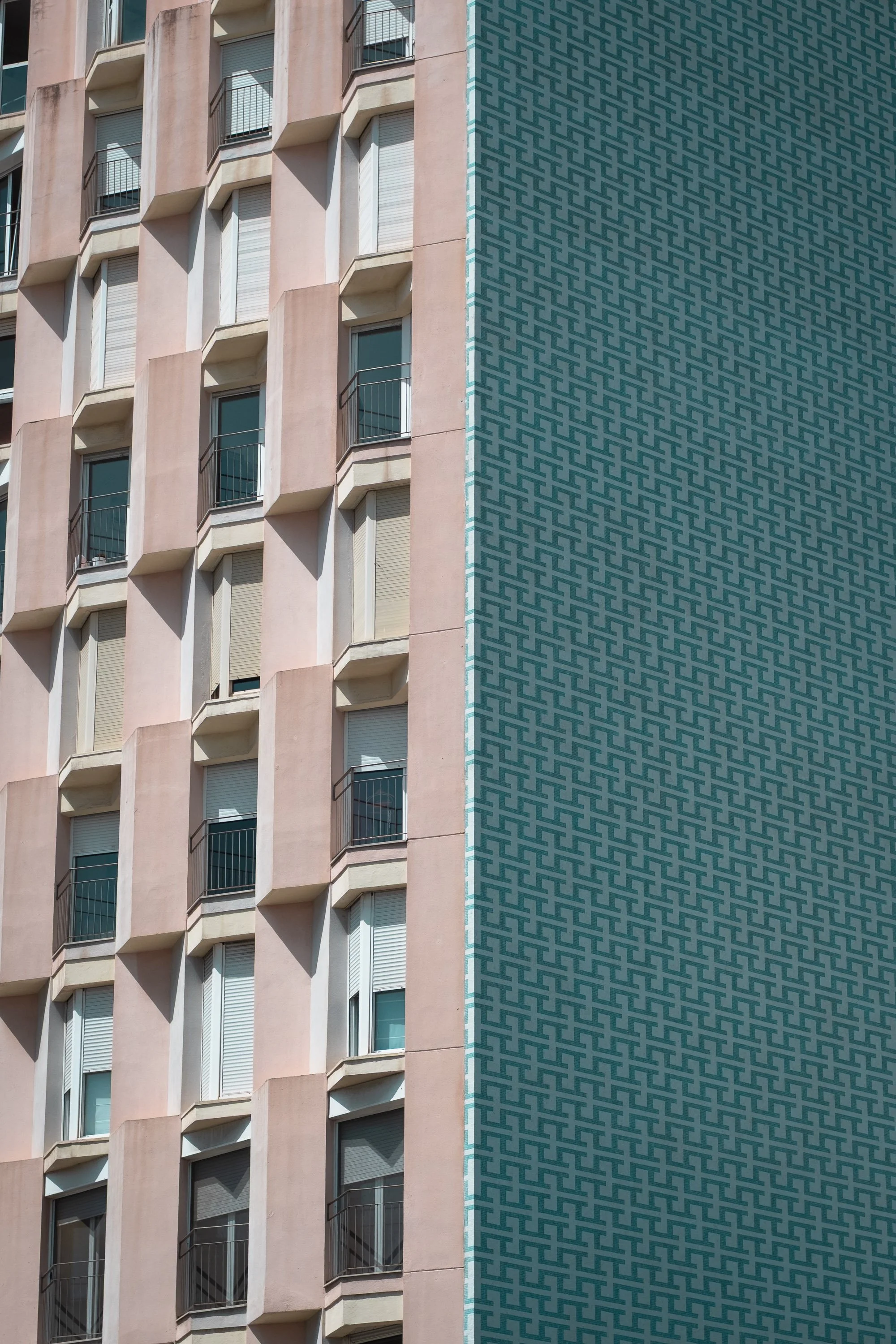 Close-up of a tall building with pink exterior walls and numerous windows, some with window shutters, and small balconies with black railings. The right side of the building features a textured teal-colored patterned section.