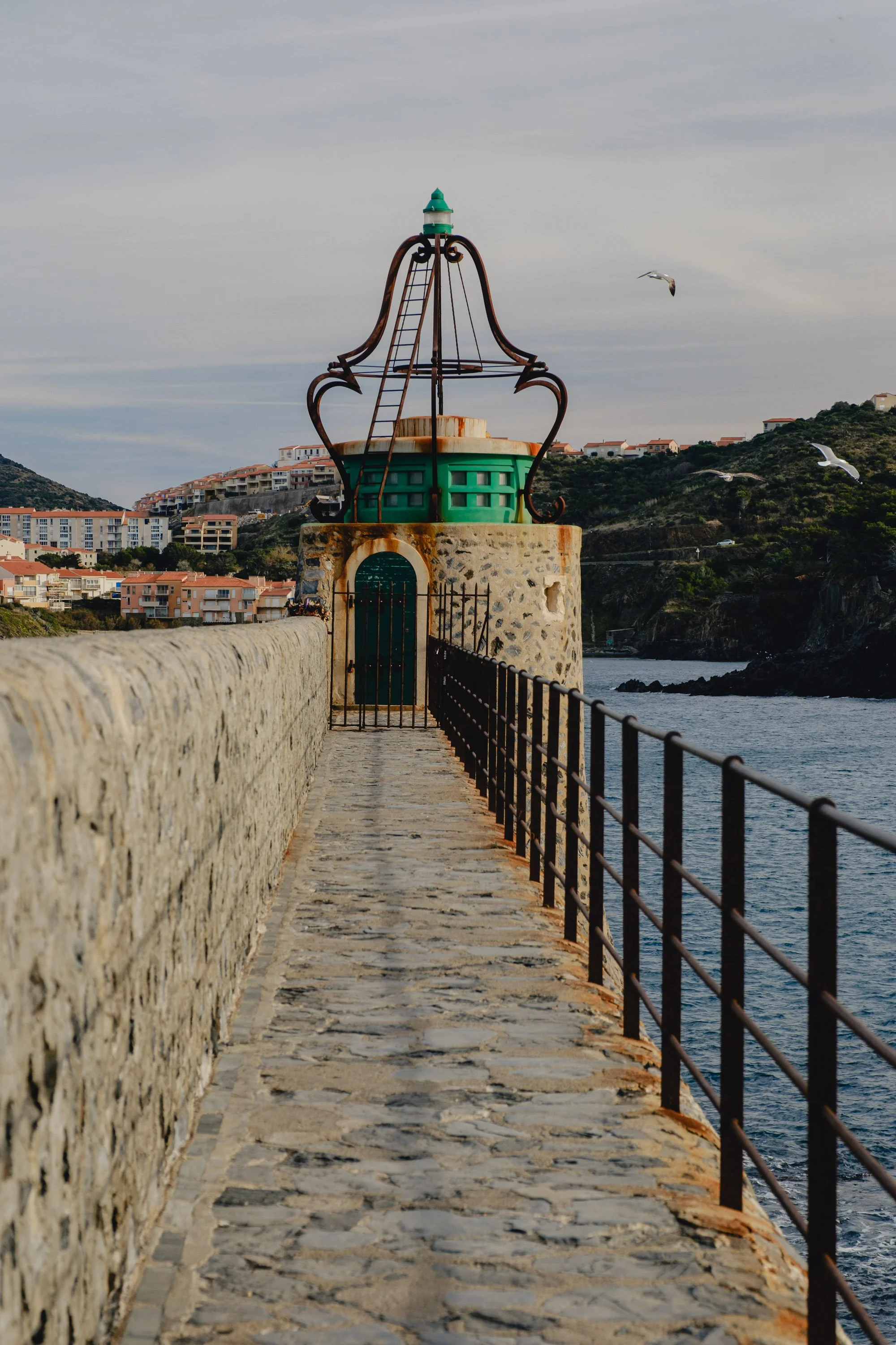 A stone pathway on a pier leading to a small lighthouse structure with a metal framework on top, overlooking a body of water with a hilly landscape and residential buildings in the background, and seagulls flying overhead.