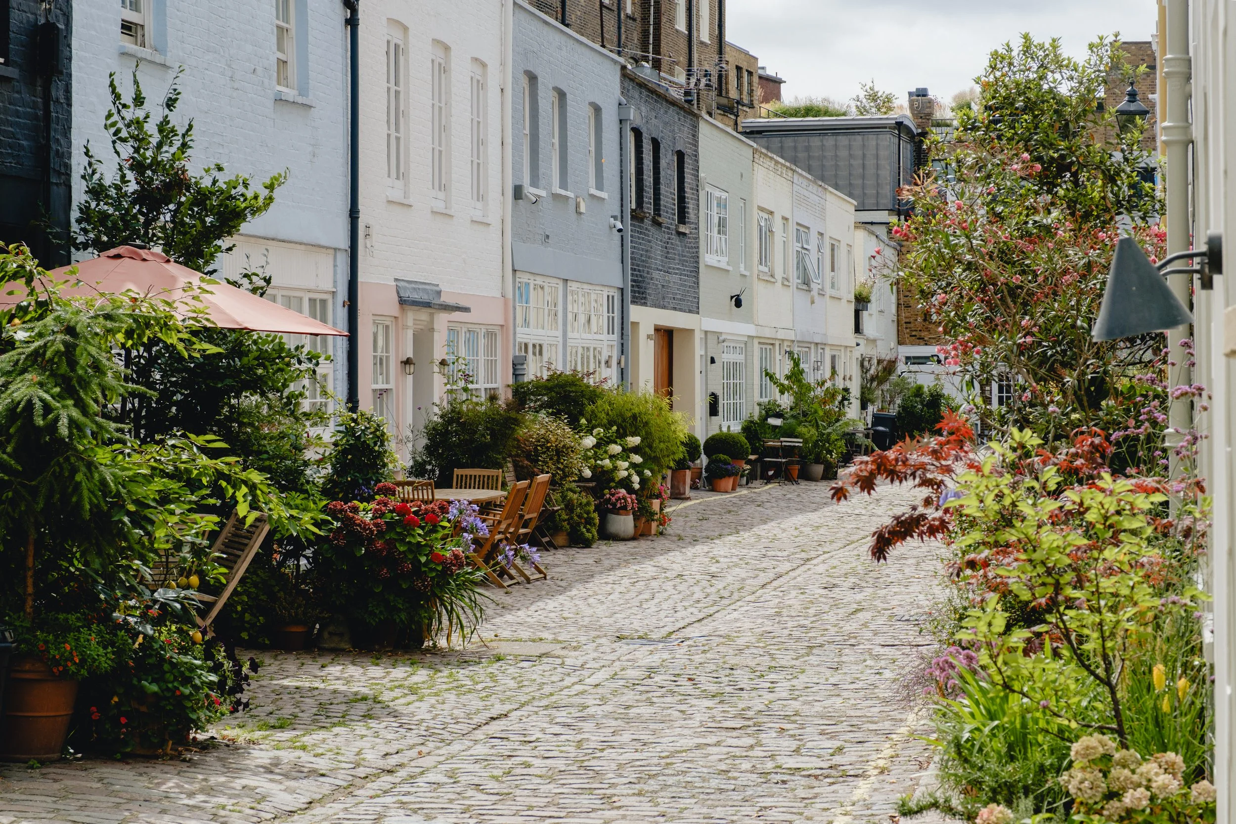 A cobblestone street lined with colorful houses and lush potted plants, with outdoor seating and pink and red flowers, under a cloudy sky.