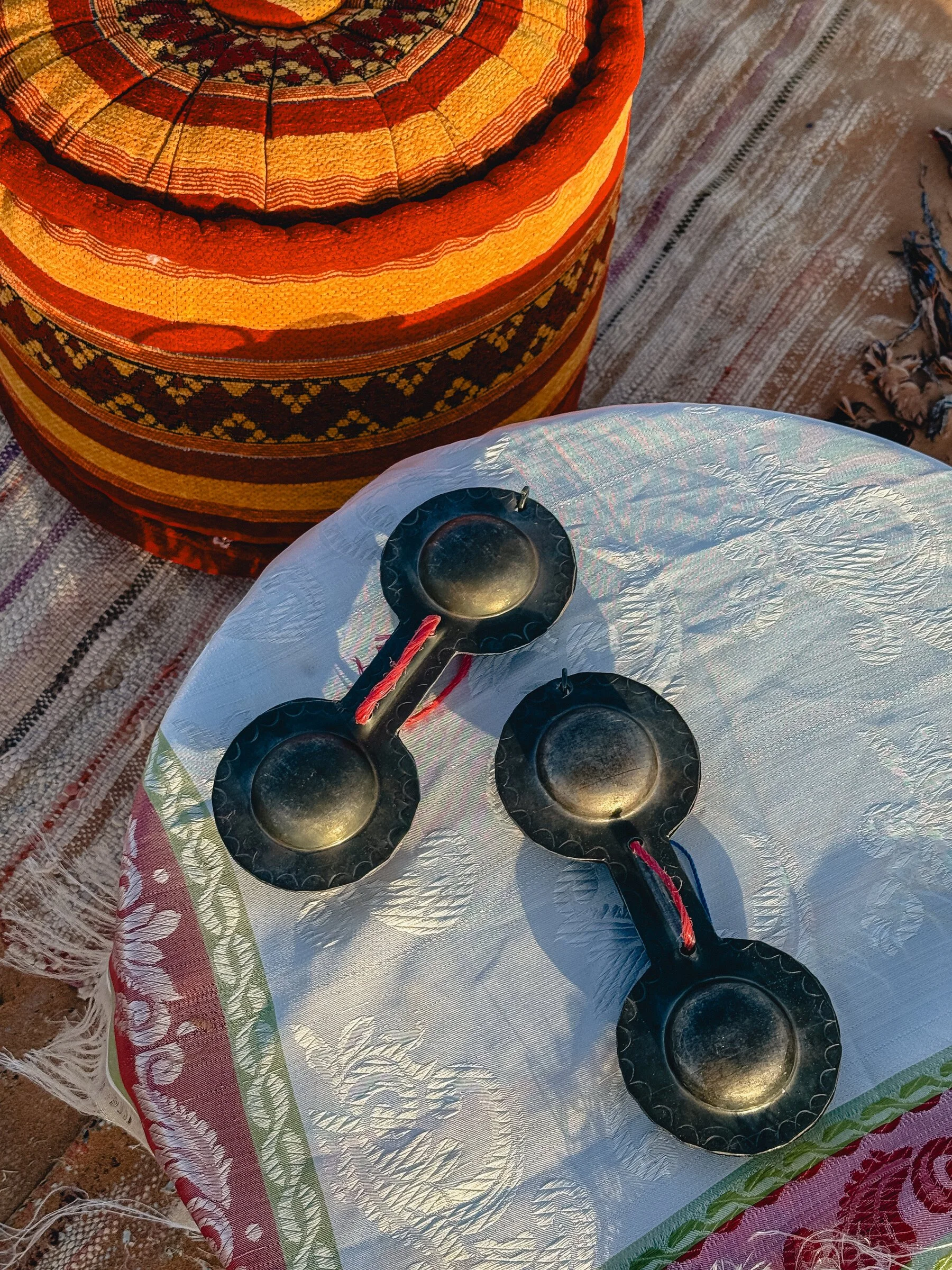 A pair of brass Gnawa castanets rests on a woven cushion beside colorful striped textiles