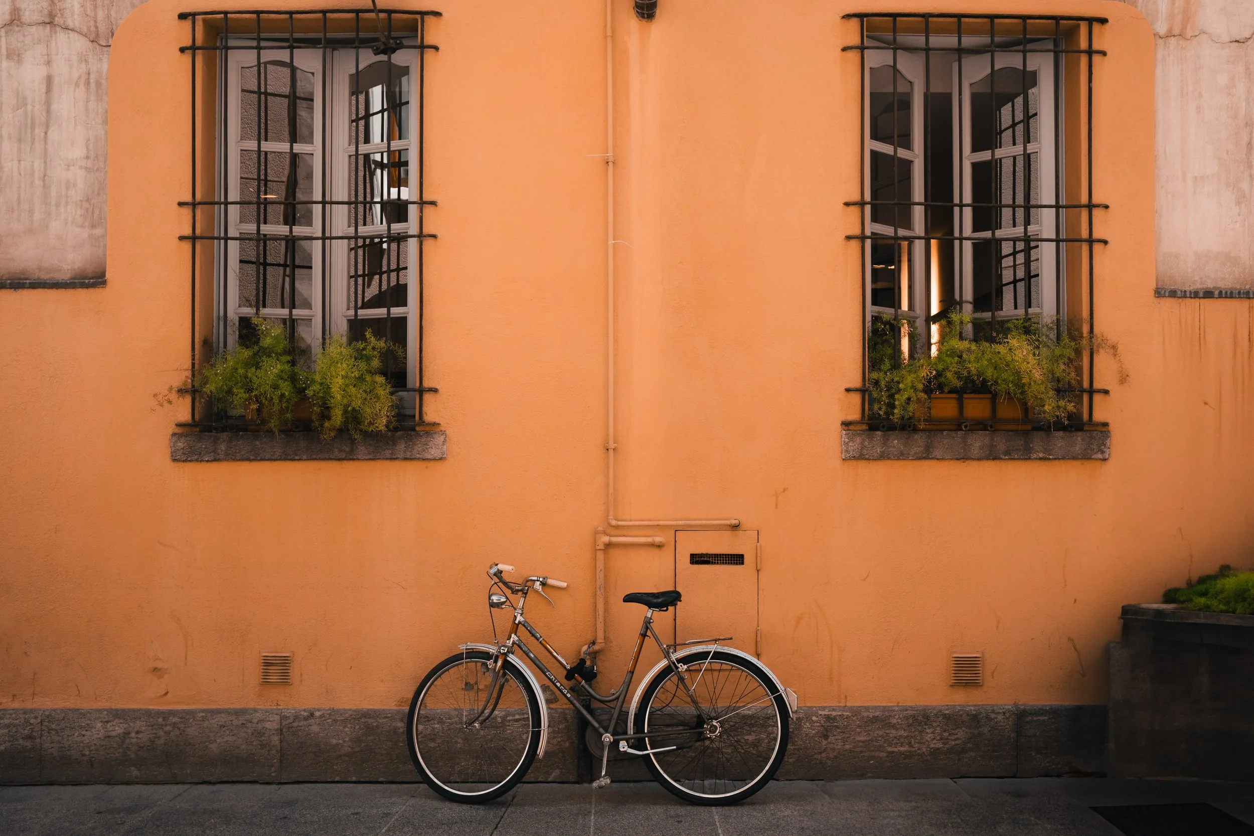 A bicycle leans against an orange building wall with two windows, each with black metal bars and flower boxes. The windows are open, revealing an interior with stairs and warm lighting.