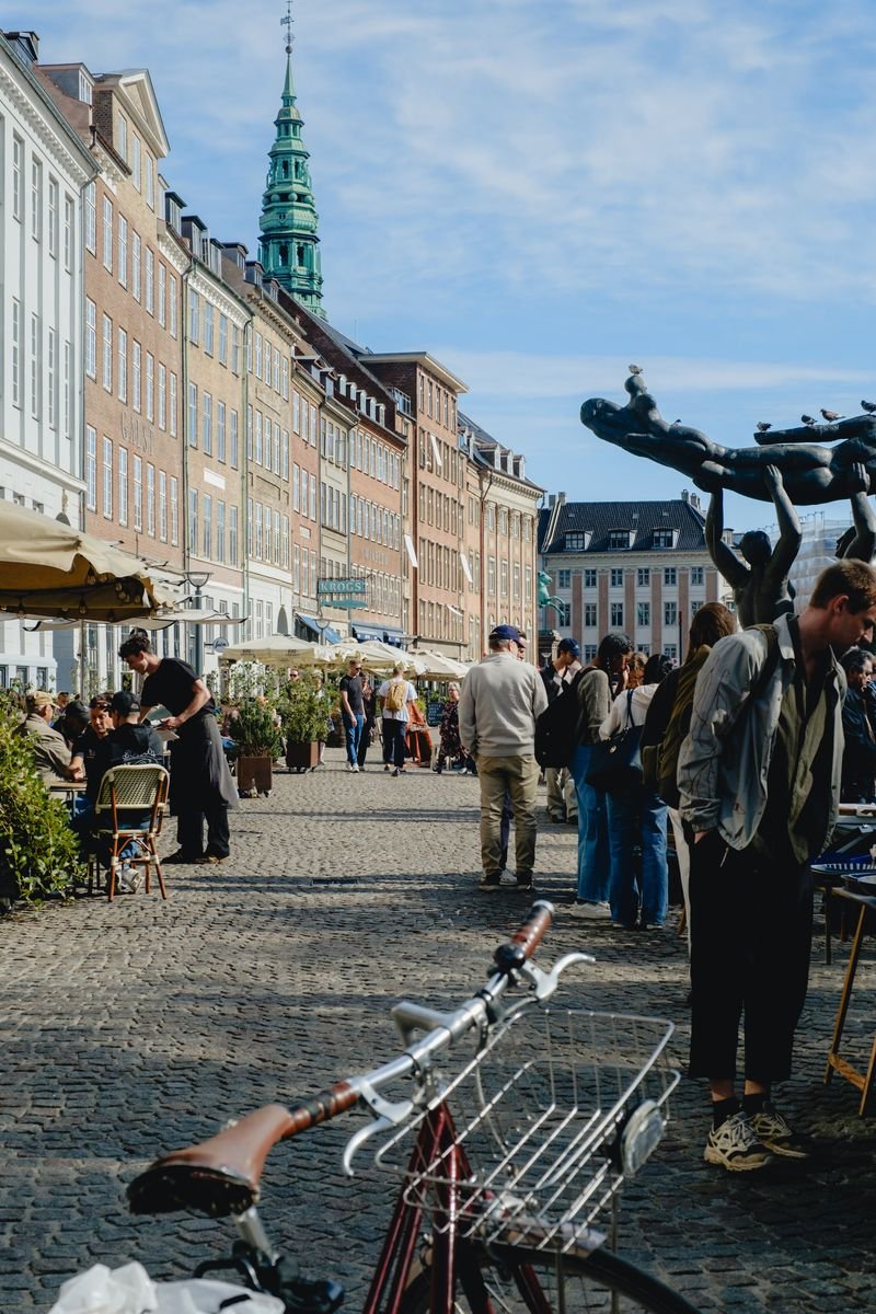 A bustling square with a cyclist, sculpture, distant spire, and cafe tables